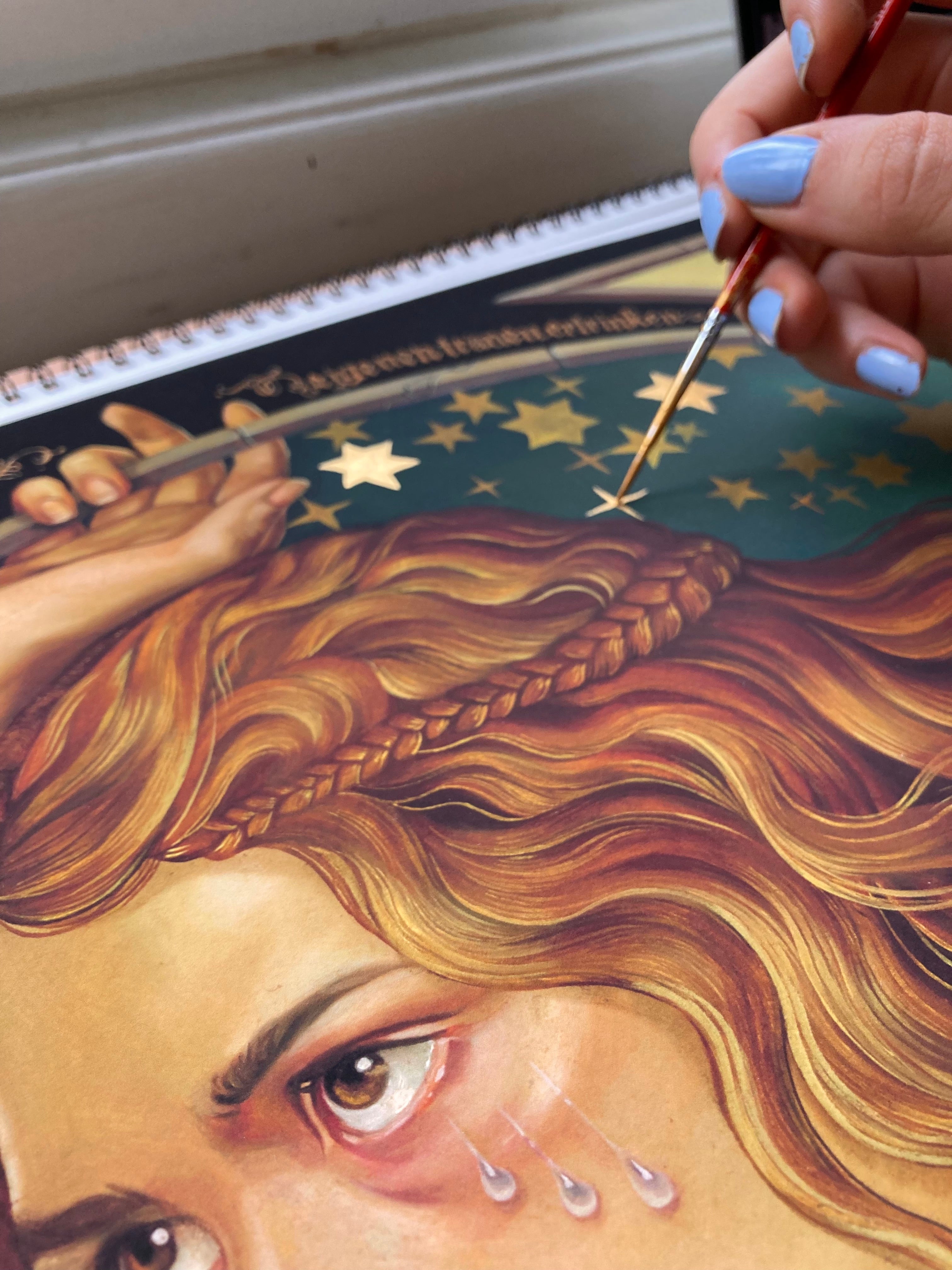 In this close-up, a person with blue-painted nails delicately touches up Alice by Ruth Speer—a hand-embellished, signed and numbered artwork—with a face featuring long hair and tears against a rich green background adorned with gold stars.