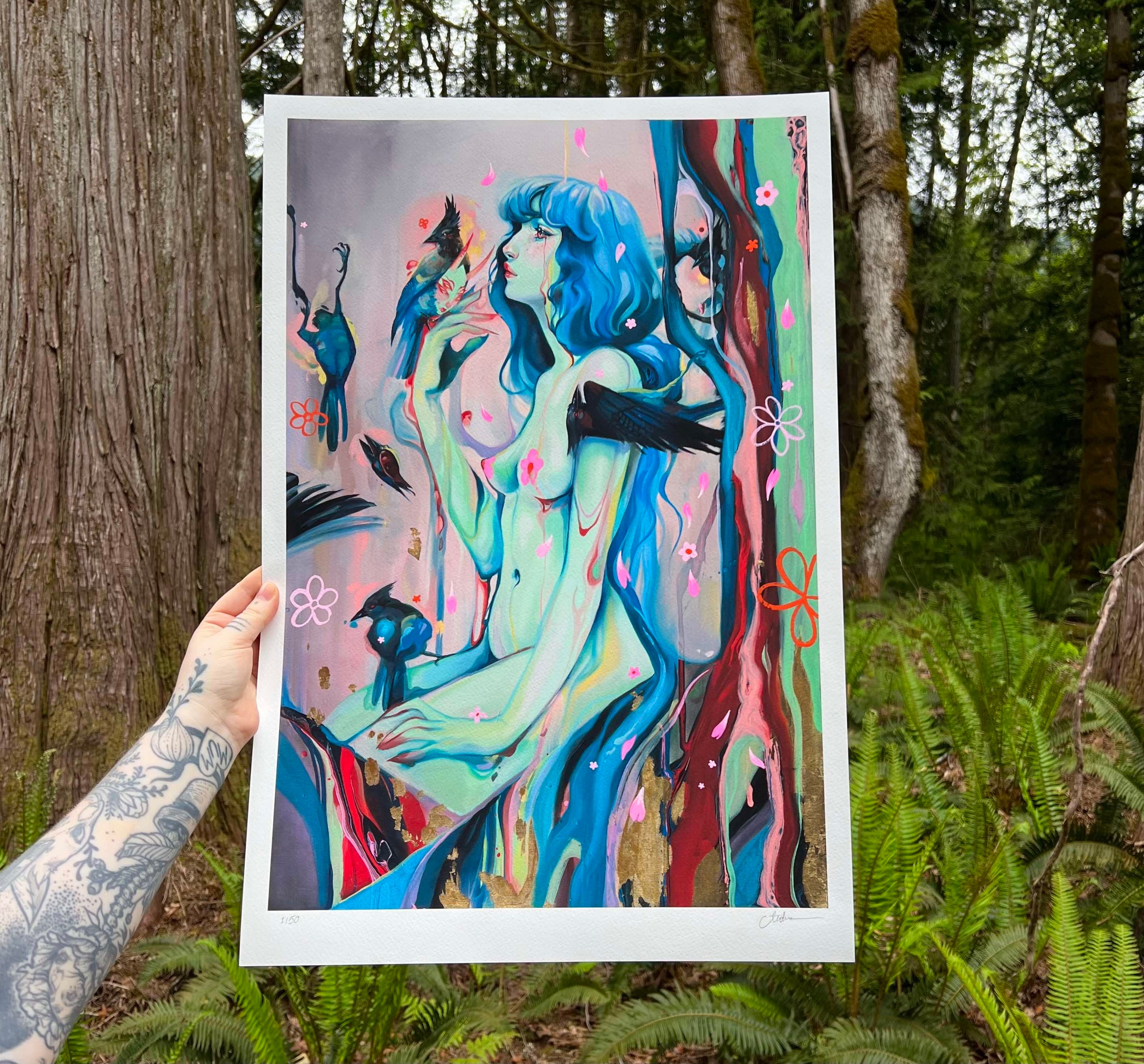 In a forest setting, a tattooed arm showcases Death & Transformation, an archival pigment print by Audra Auclair. The hand-embellished artwork features a blue-haired figure surrounded by birds and abstract shapes, merging surreal elements with nature amid tall trees and greenery.