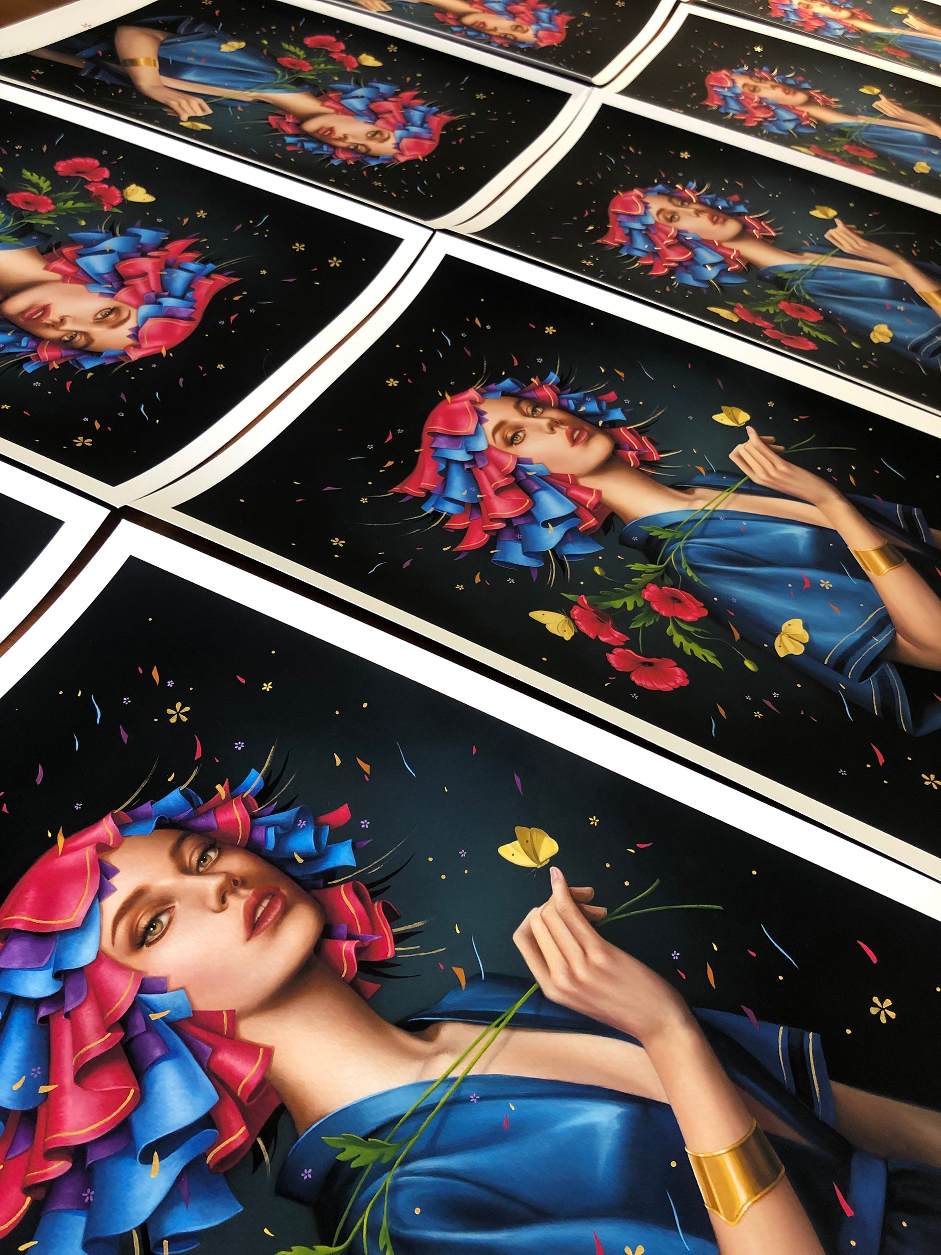 The Poppy series by Allison Reimold features signed, numbered prints of a woman with pink and blue flowers in her hair, holding a yellow flower. She wears a blue garment and gold bracelet against a dark background with confetti details, each print hand embellished for added allure.