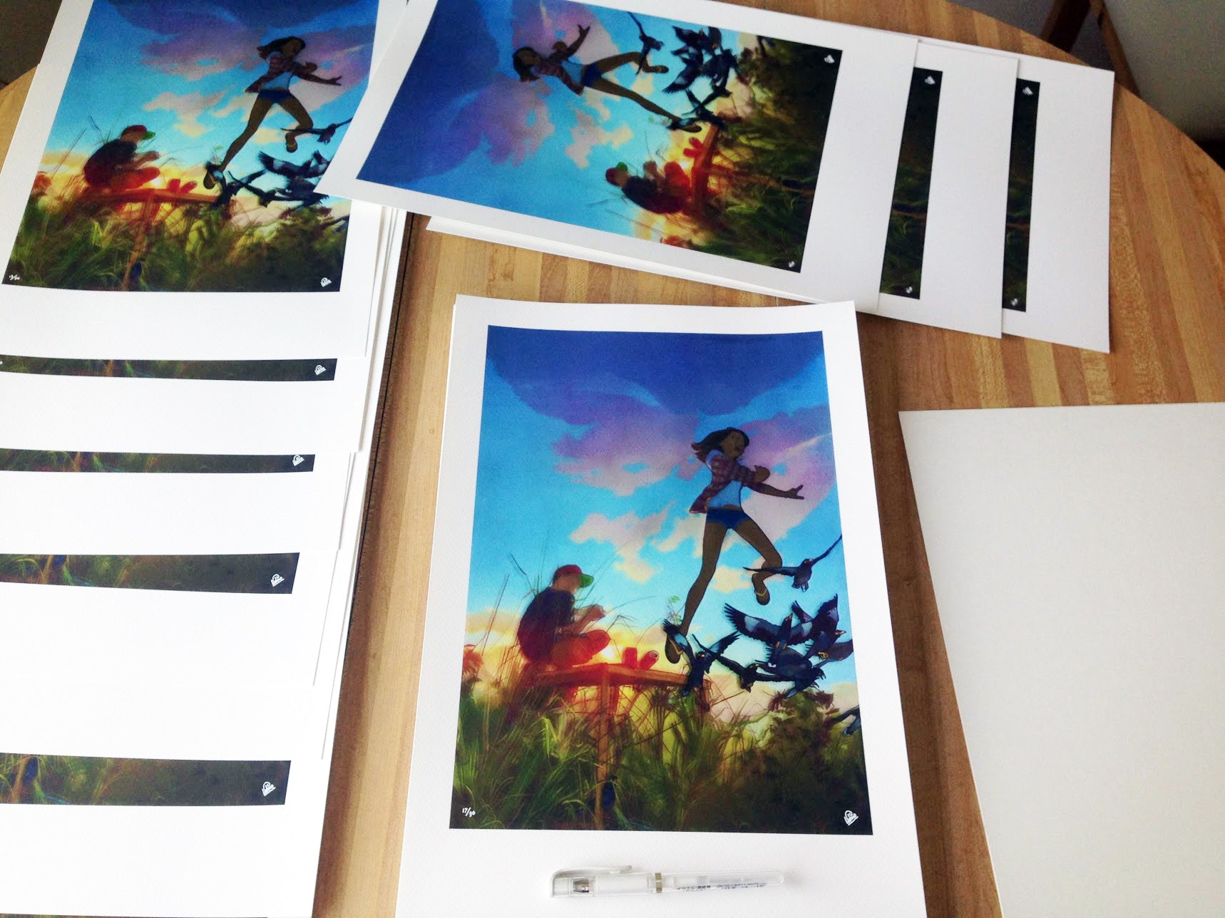 Edwin Ushiros vivid artwork, titled One More Second You Are About to Gain, features people jumping in a scenic outdoor setting. These archival pigment prints are spread across a wooden table, with a pen nearby ready for signing and numbering.