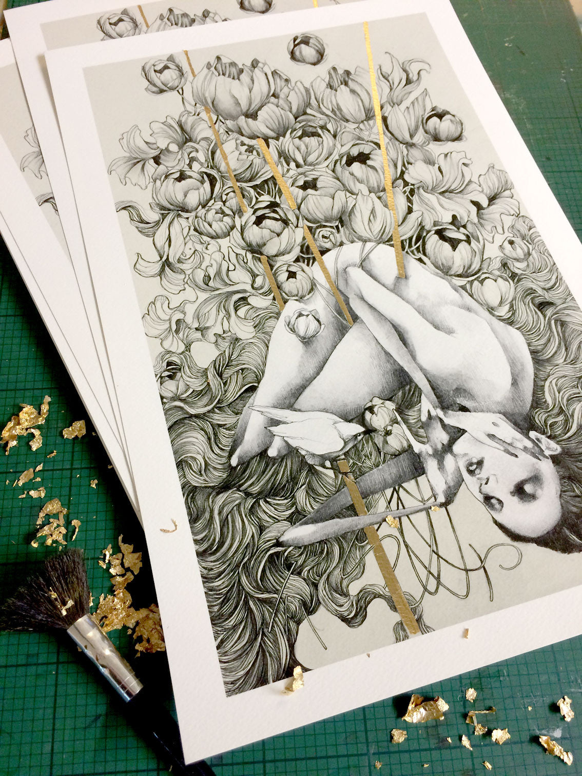 The Embrace fine art giclee prints by Craww depict a person amid flowers and flowing hair, highlighted with hand-embellished gold leaf accents. A paintbrush and gold leaf flakes rest nearby on the green surface.