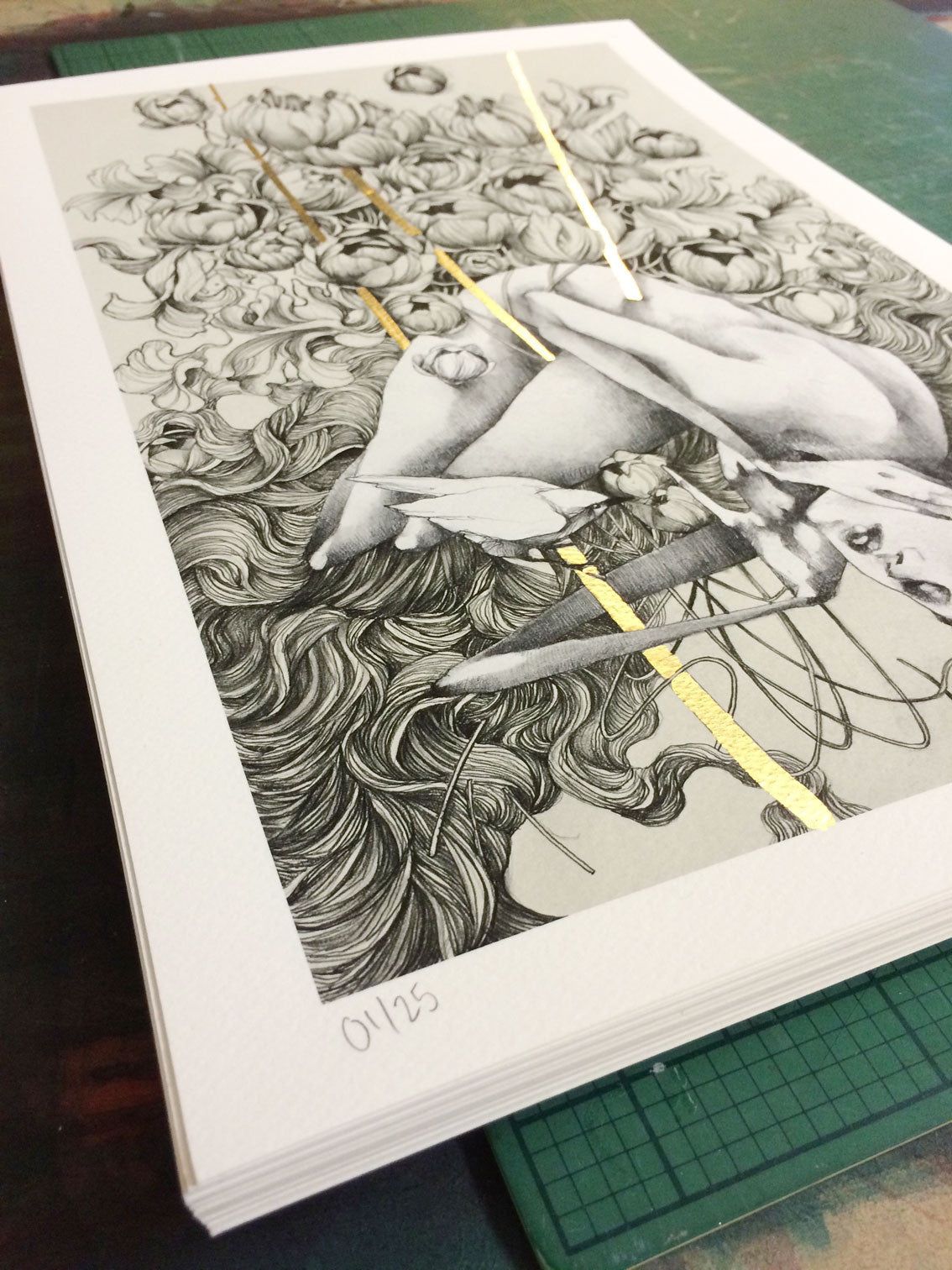 A stack of Embrace black and white fine art giclee prints by Craww features swirling patterns, a figure, and gold leaf accents. The bottom print is marked 01/25, the first in this hand-embellished limited edition series.