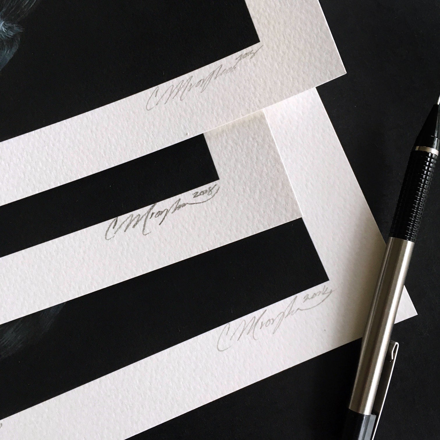 Close-up of three Limited Edition Christina Mrozik art prints titled We Will Never Get To The Bottom Of It, each signed Moyson 2018, on a dark surface. A pen with a silver tip and black grip lies beside the textured edges of the Archival Pigment Prints.