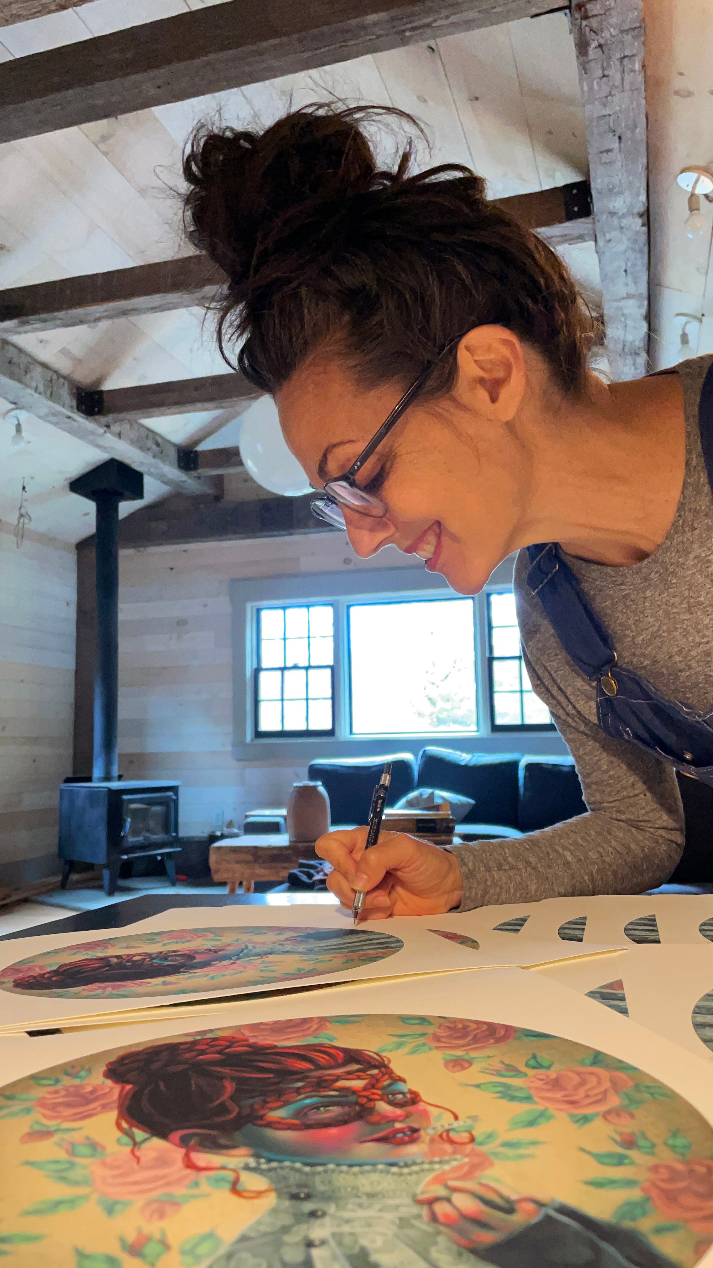 A woman with glasses and a bun smiles as she signs Profanatine prints by Lori Nelson in a cozy room with wooden beams and a stove. Each colorful archival pigment print, from an edition of 40, is personally signed and numbered, enhancing their unique charm.