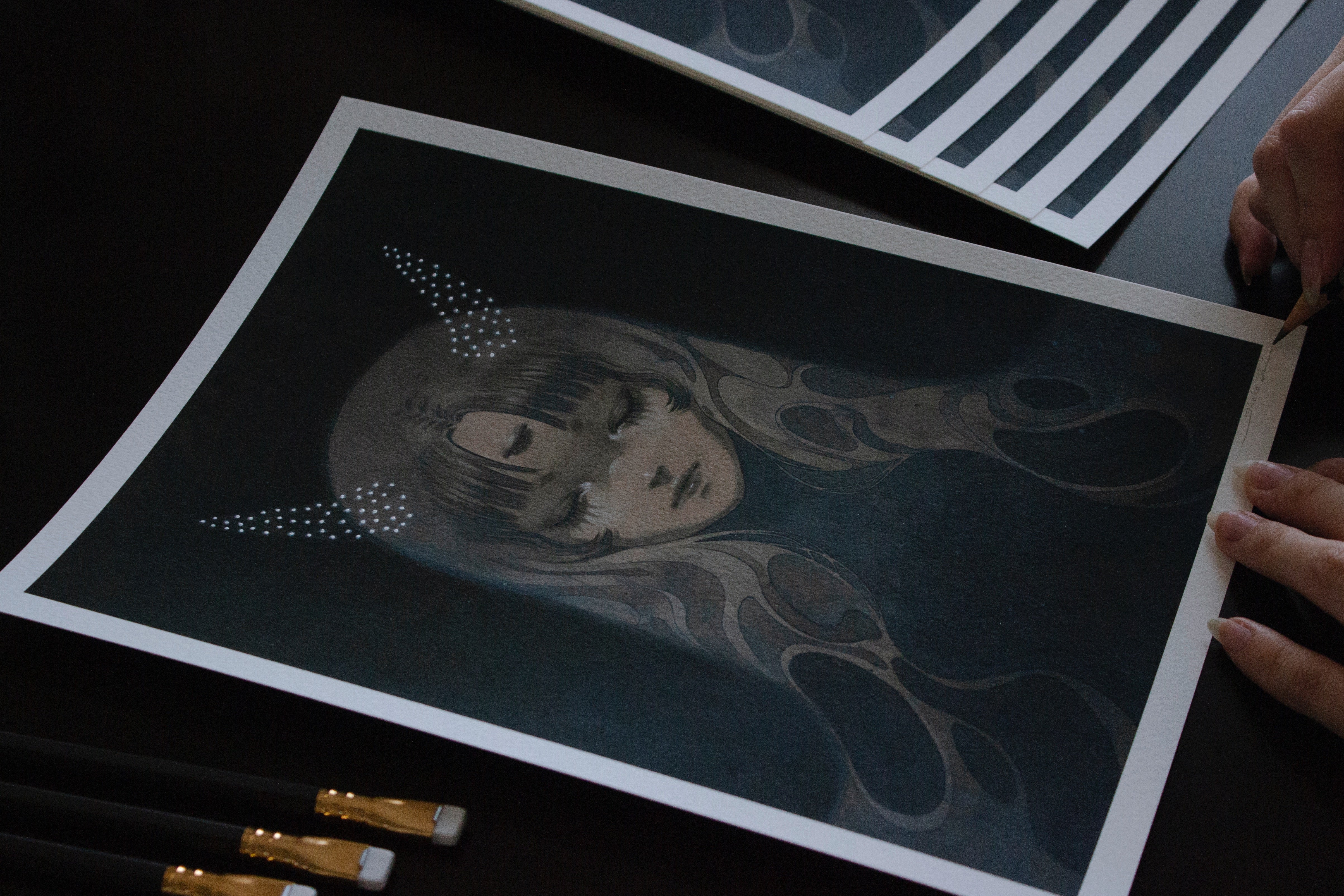 An artist is crafting the dark, stylized portrait Stardust Slumber, depicting a woman with closed eyes and animal-like ears. Several paintbrushes lie nearby, emanating Shoko Ishidas mysterious, ethereal style.