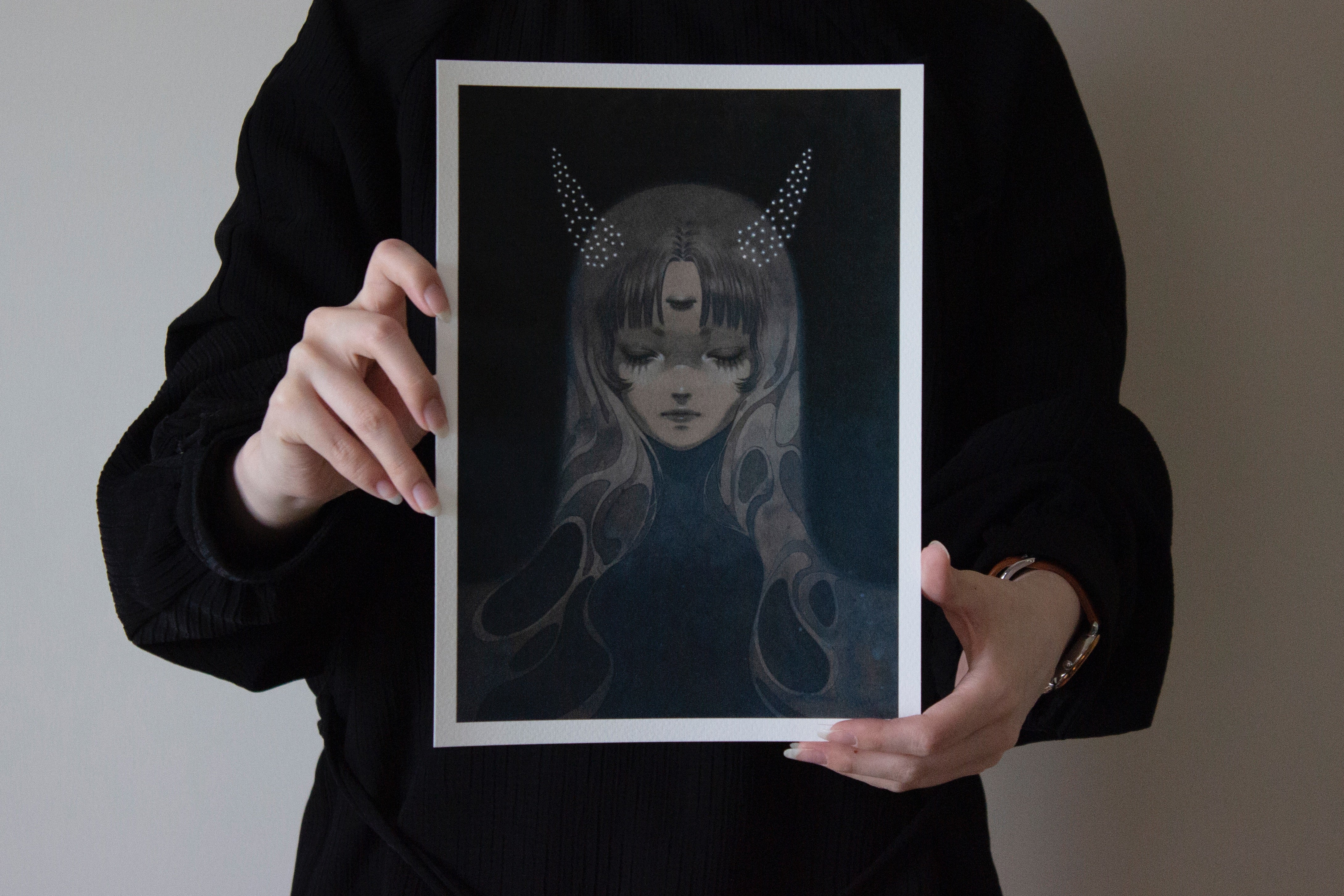 A person in black holds Shoko Ishidas archival pigment print Stardust Slumber, featuring a dark, artistic image of a female figure with closed eyes, a third eye on her forehead, hair flowing like stardust, and dotted horns against a plain background.