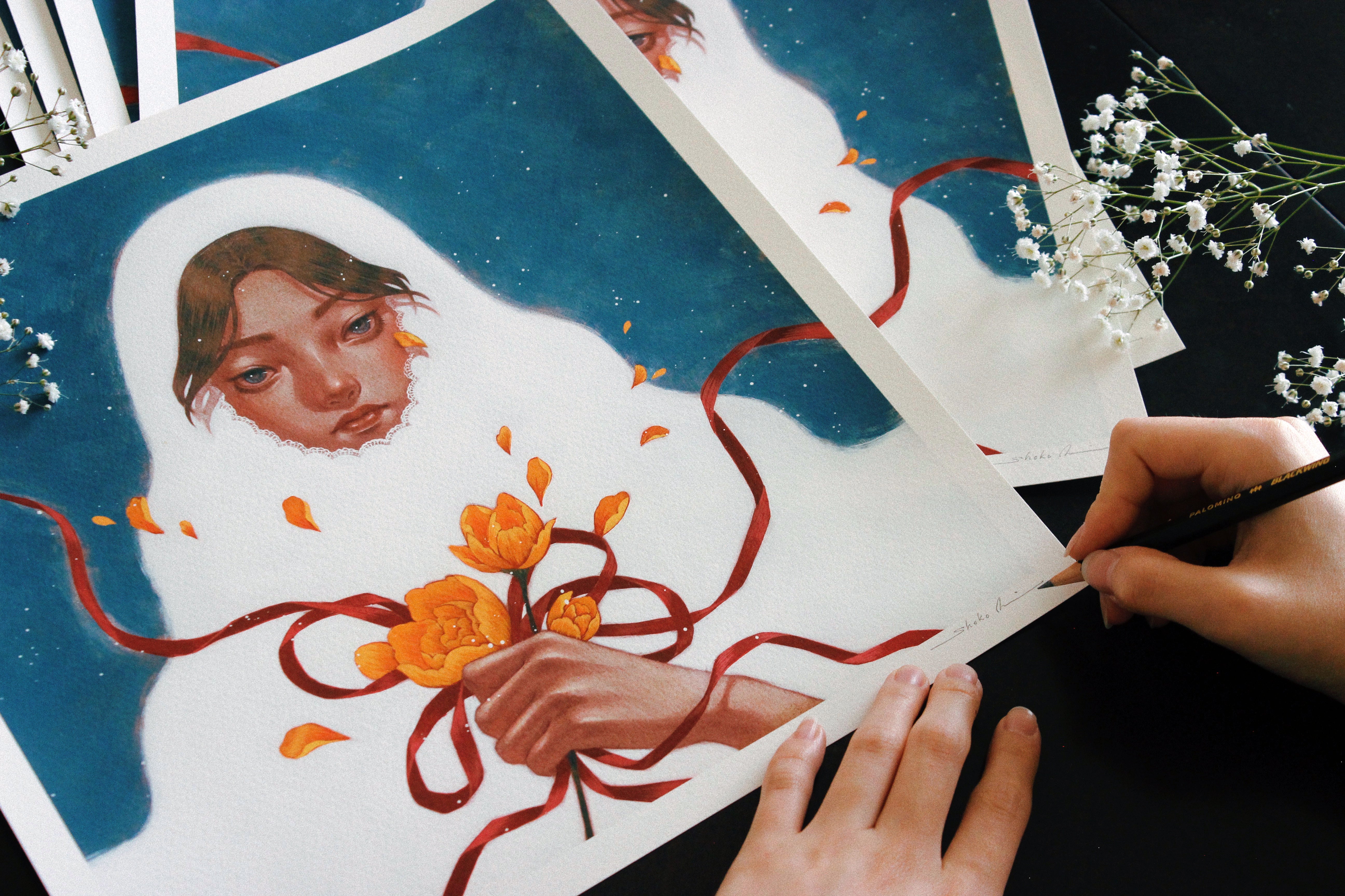 The artist Shoko Ishida signs an archival pigment print titled Winter Bride, featuring a figure in a white shawl holding orange flowers with a red ribbon, set against a blue backdrop, with delicate white flowers on a dark surface.