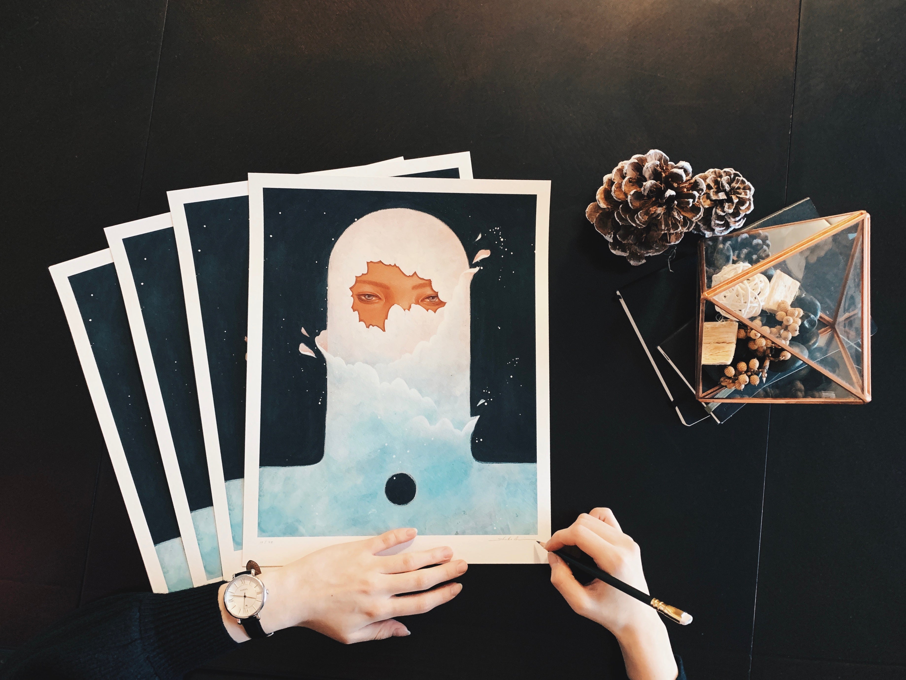 A person wearing a watch is signing and numbering archival pigment prints titled In Daydream by Shoko Ishida. The prints display a surreal face rising from clouds, with pine cones and a geometric container filled with nuts on the dark table.