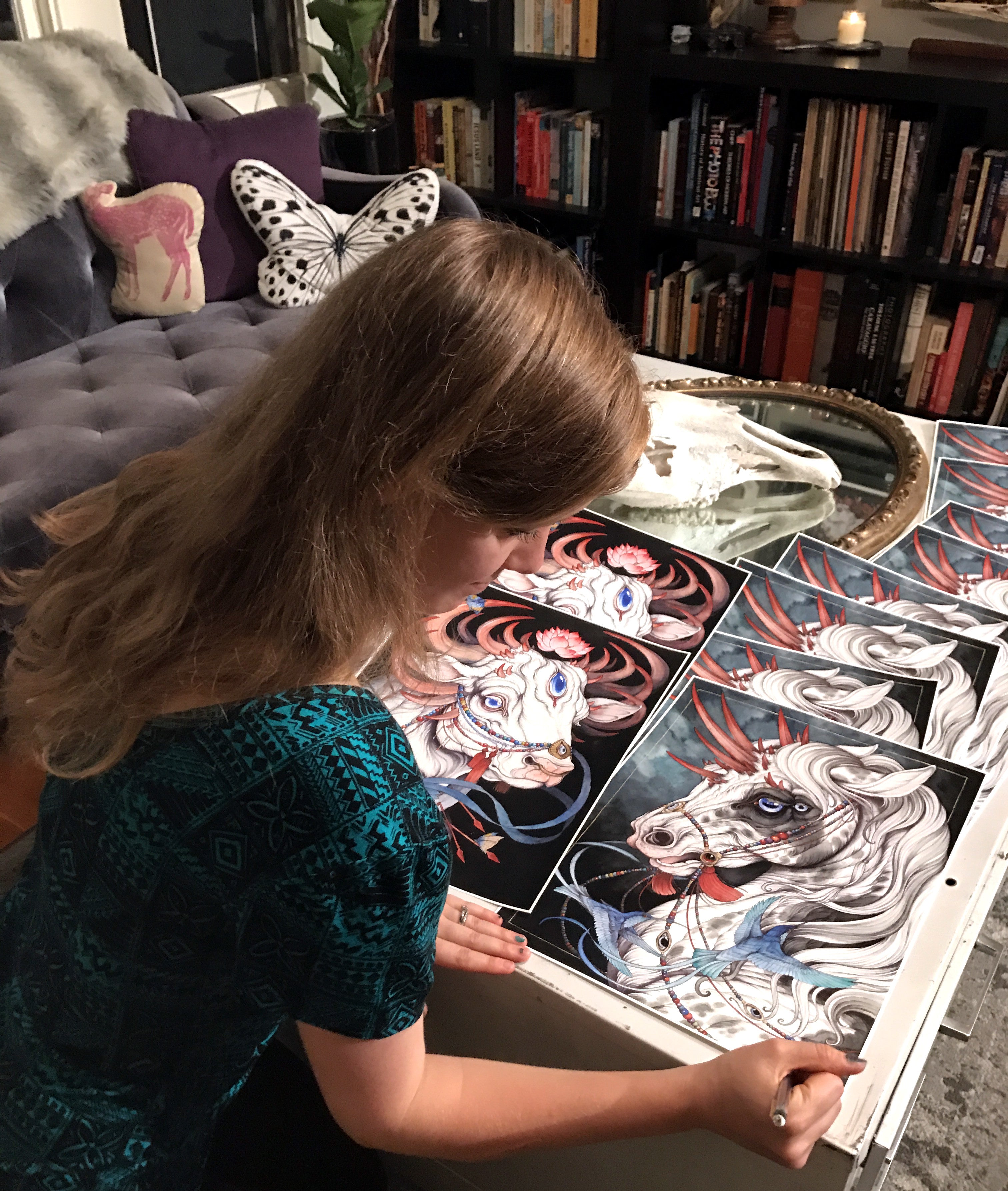 In a cozy, book-filled room adorned with deer-themed decor and an animal skull, Caitlin Hackett, wearing a teal patterned shirt, sits at a table. Her long hair cascades as she meticulously illustrates The Gift Horse, to be recreated as an archival pigment print.