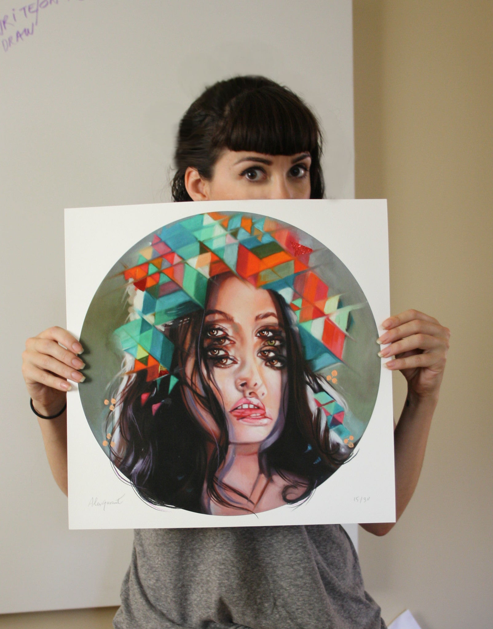 A person with dark hair and a gray shirt displays Kaleidoscope, an artful fine art giclee print by Alex Garant, featuring a woman embellished with geometric shapes and multiple eyes. The plain wall behind is partially obscured by the vibrant artwork.