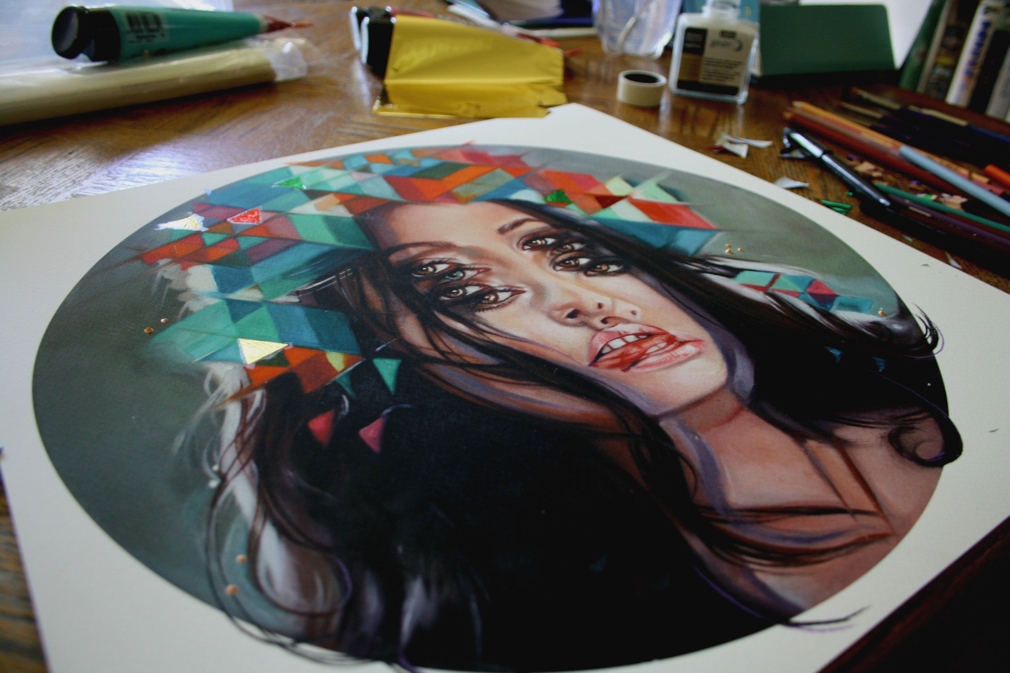 The Kaleidoscope by Alex Garant is a surreal painting of a womans face with overlapping eyes and vivid geometric shapes, resembling a kaleidoscope. Its surrounded by art supplies on a wooden table, making it perfect for a fine art giclee print.
