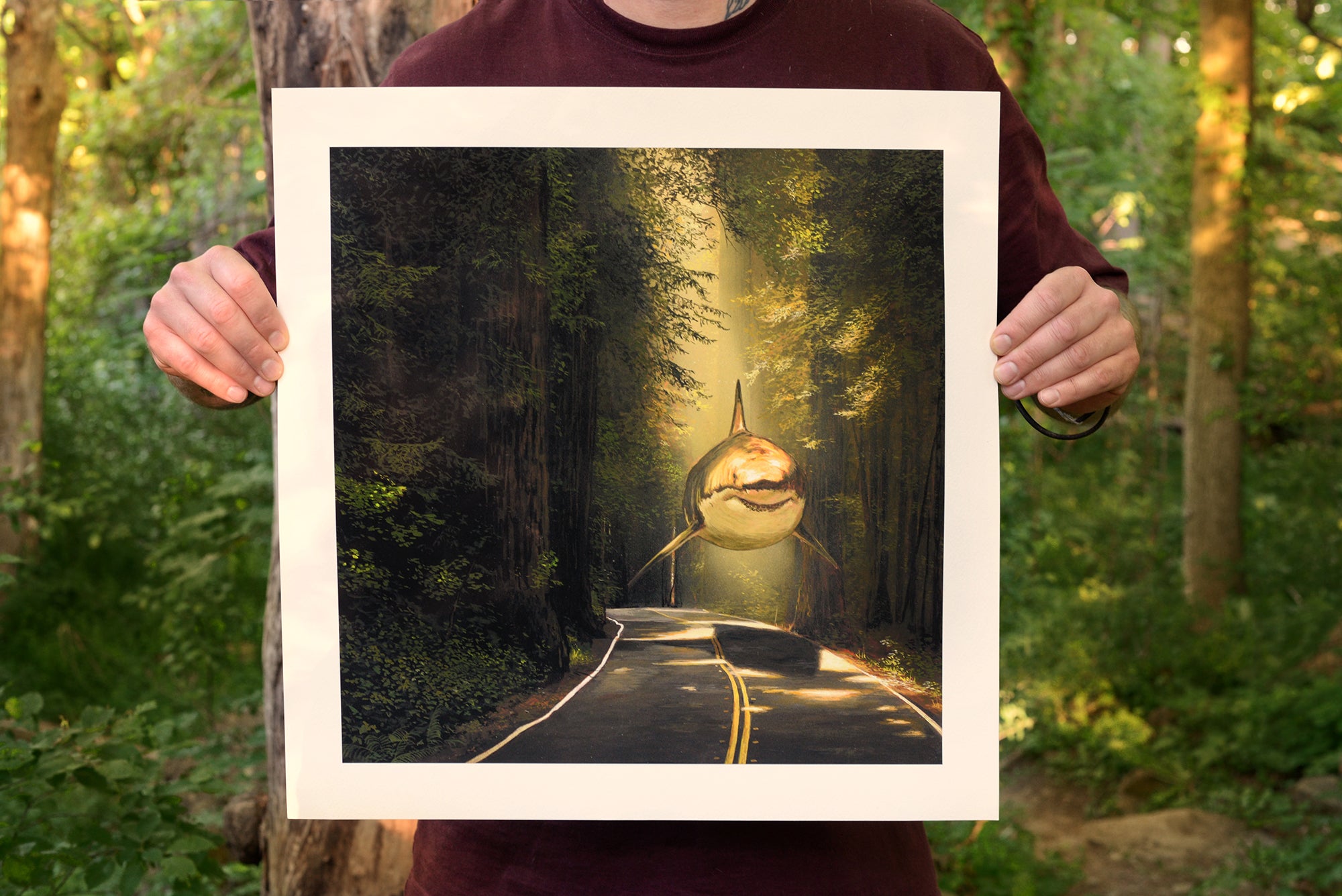 A person holds Chris Austins Rewarded Are The Curious, a limited edition framed archival pigment print. It depicts a surreal scene of a shark swimming down a sunlit forest road, contrasting the dense tall trees and sea creature. Each piece is signed and numbered.