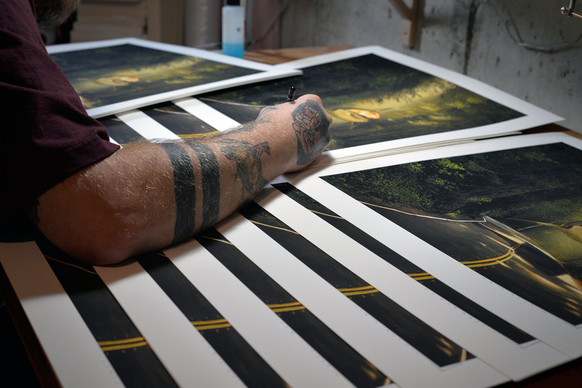 Chris Austin, a tattooed artist, signs and numbers limited edition prints of the Rewarded Are The Curious painting. These archival pigment prints depict a sunlit road with tree-lined landscapes and are carefully handled while arranged neatly on a table.