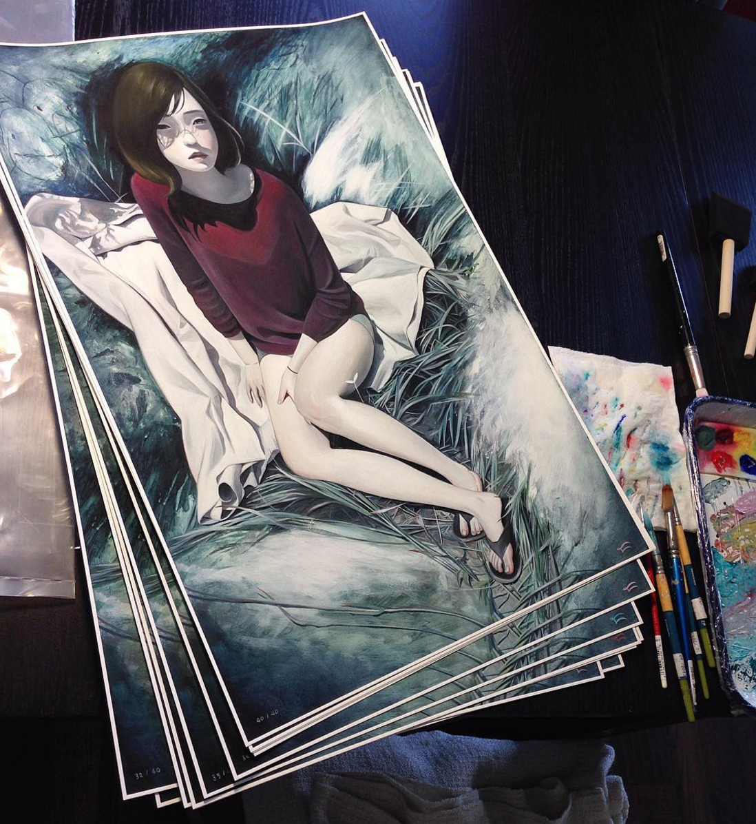 The Limited Edition Lucid fine art giclee prints by Joanne Nam depict a woman in a burgundy top and white skirt lying on grass, accompanied by art supplies like brushes and pencils, capturing the elegance of her signature style.