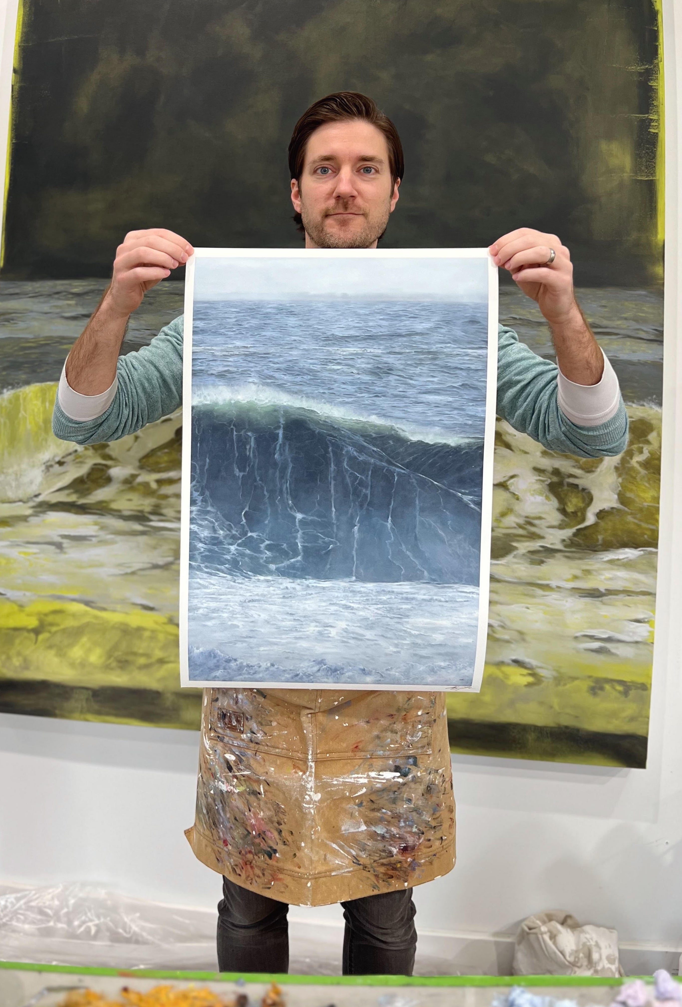 A person in a light blue sweater and paint-splattered apron holds an archival pigment print of a wave similar to Goliath Dreams by Adam Hall, with another ocean-themed painting in the background. Art supplies are visible at the bottom.