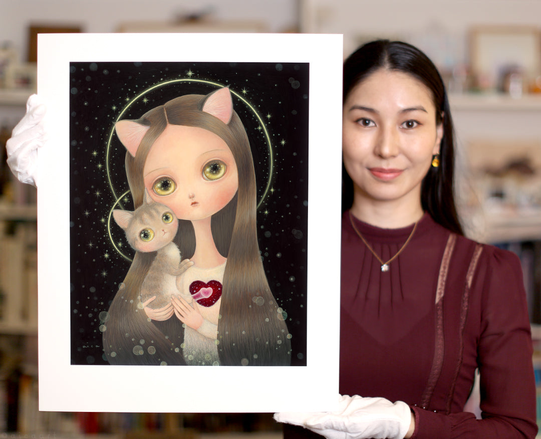 A woman in a burgundy dress and white gloves displays "Heart" by aica—an archival pigment print featuring a girl with cat ears and long hair holding a cat, both with large eyes, set against a dark starry background.