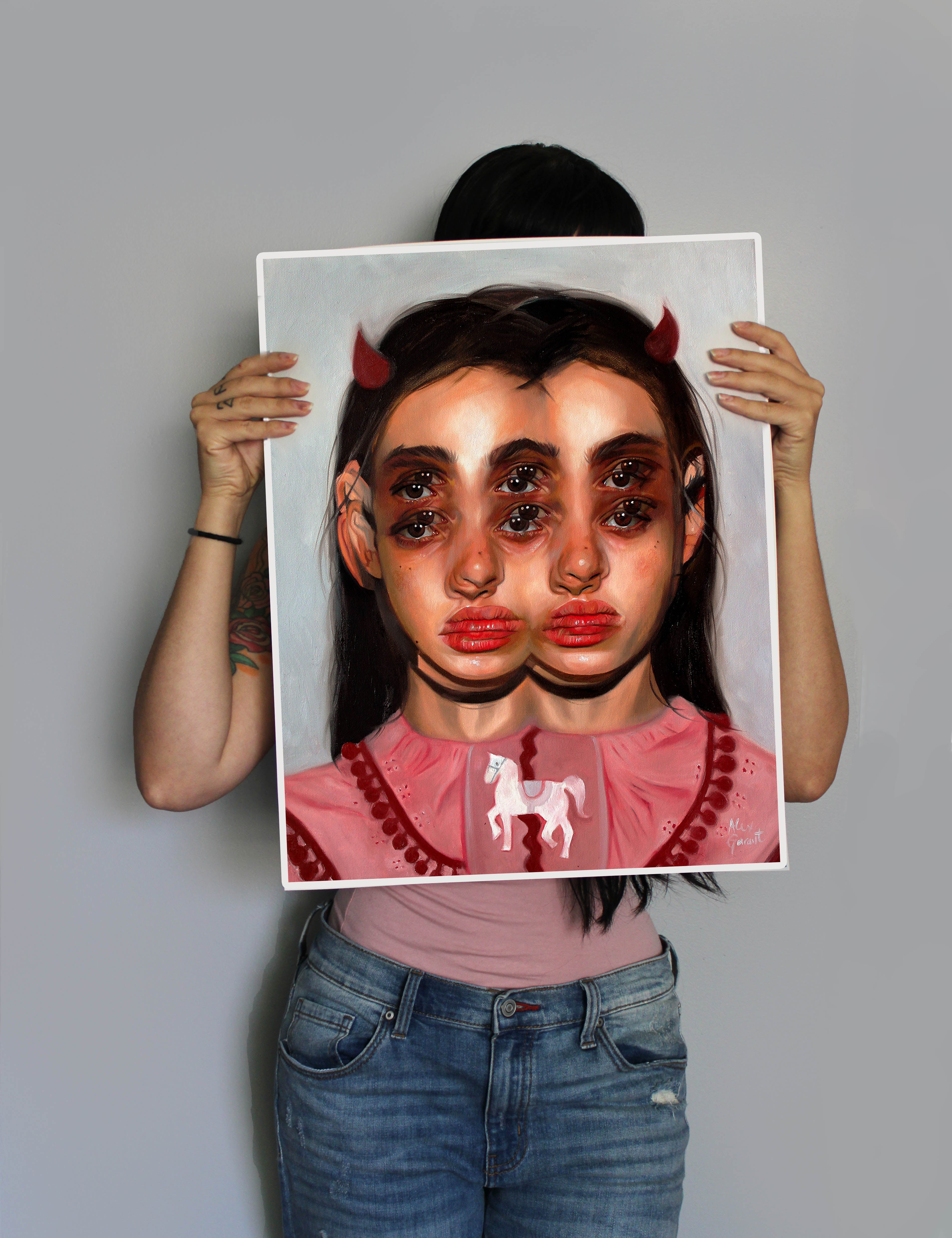 A person holding Carousel, an Alex Garant surreal painting, obscures their face. The artwork shows a girl with overlapping faces, red horns, and a unicorn on her pink shirt. Their arms in jeans and a dark top peek out from behind the mesmerizing archival pigment print.