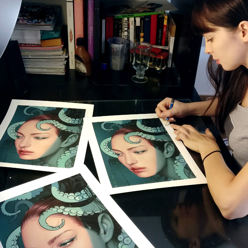A woman at a desk meticulously enhances Cecaelia by Sarah Joncas, featuring a womans face entwined with tentacles like a cecaelia. Books and archival pigment prints are scattered among her art supplies in the background.