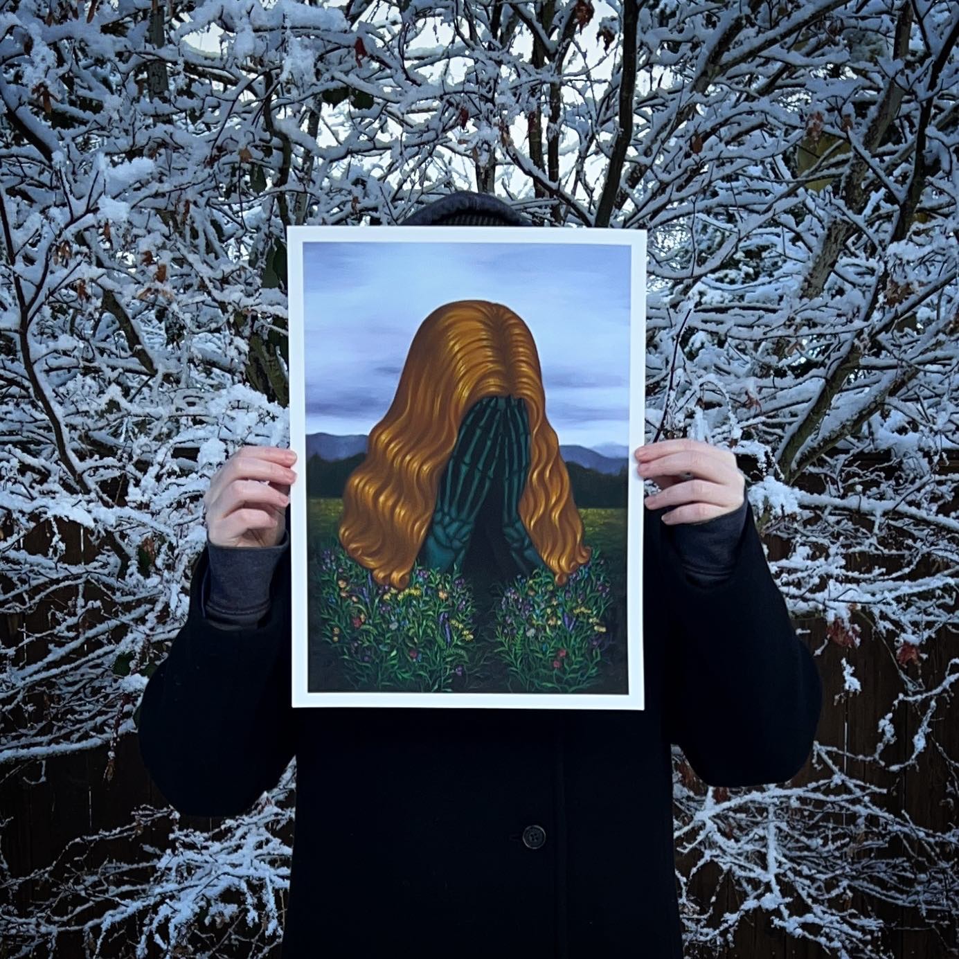 A person in a dark coat holds up Mistress of Grief, an archival pigment print by Syd Bee, featuring a wavy-haired figure hiding their face with snow-covered branches adding contrast to the colorful art.