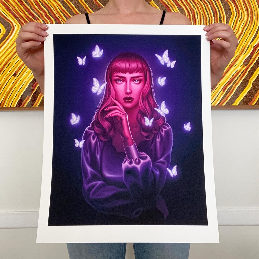 A person holds The Ultraviolet Gardener, an archival pigment print by Allison Reimold. It depicts a woman with pink hair in a purple blouse, surrounded by glowing butterflies on a dark background, contrasting vibrantly against the colorful abstract wall behind.