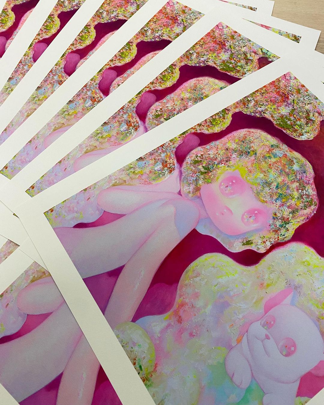 The Sanctuary limited edition archival pigment print by So Youn Lee features a stylized figure with curly hair and a small animal in a whimsical composition of vivid pinks, greens, and golds. Multiple prints are elegantly fanned out on a wooden surface.