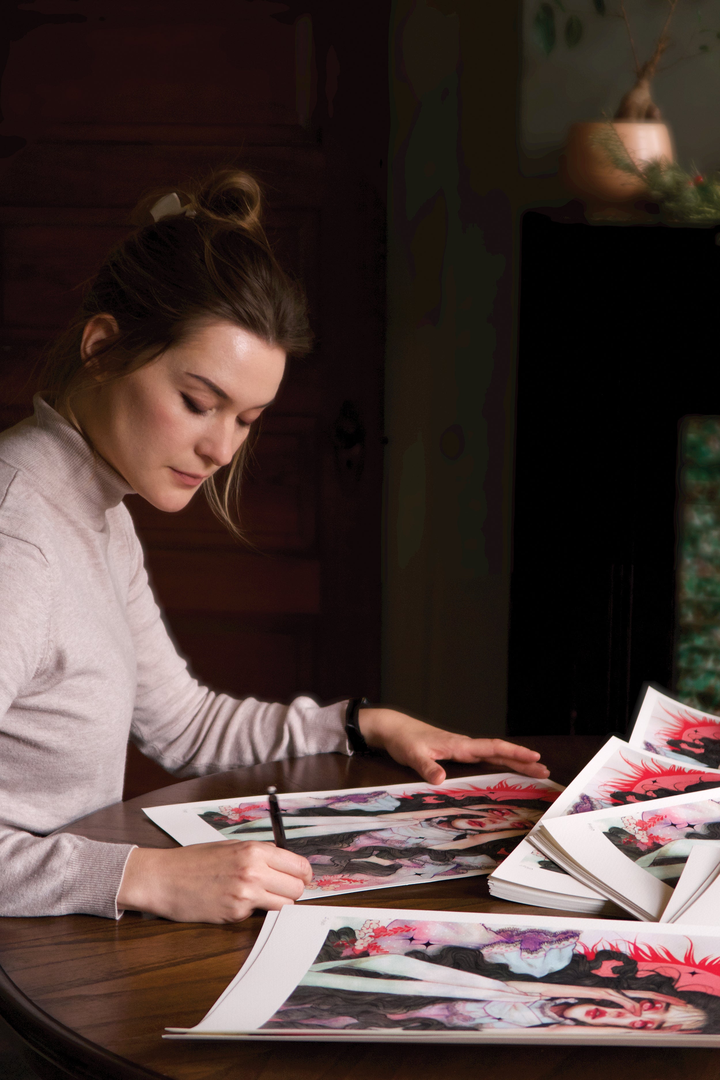 A woman with tied-back hair is seated at a wooden table, intently signing limited edition archival pigment prints of Dread by Kelsey Beckett. In front of her is a stack of these prints. The softly lit room fosters a warm and calm ambiance.