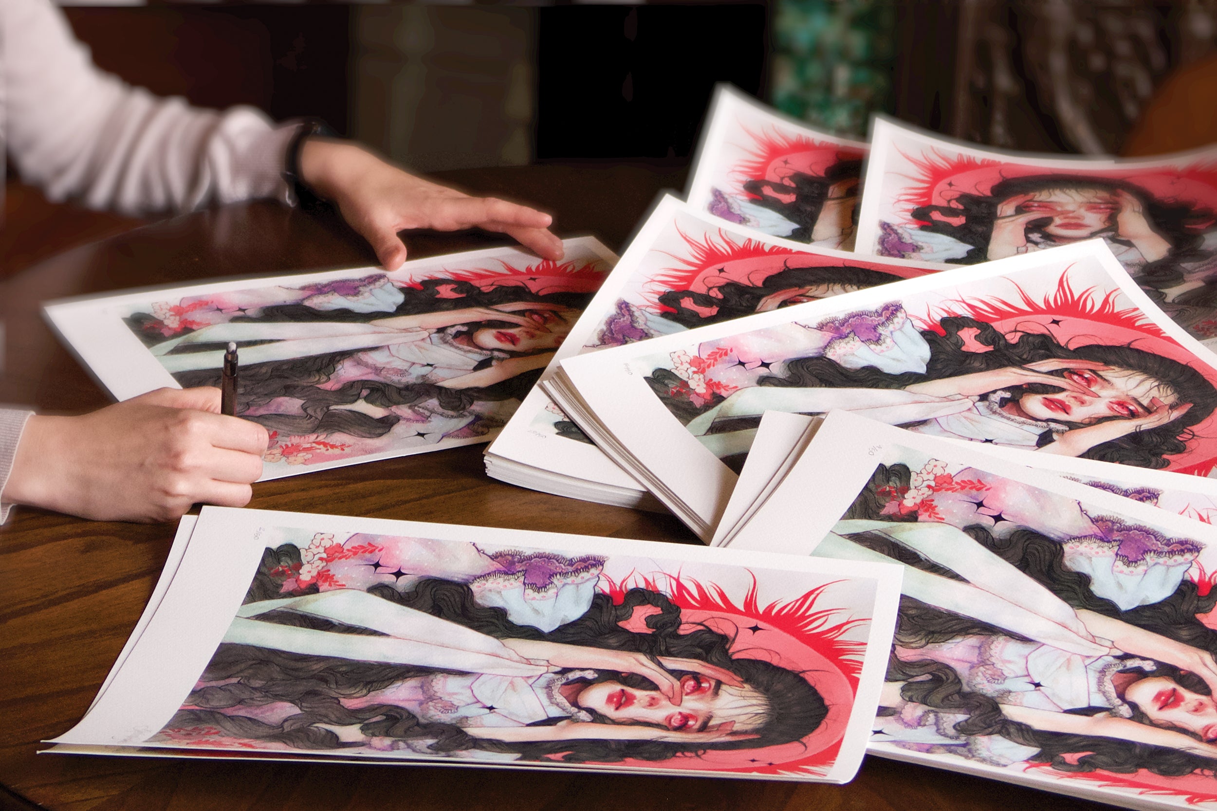 Kelsey Beckett is hand-signing several copies of her limited edition archival pigment print, Dread, featuring a vibrant illustration of a woman in vivid reds. The dramatic and intricate artwork is spread across a wooden table.
