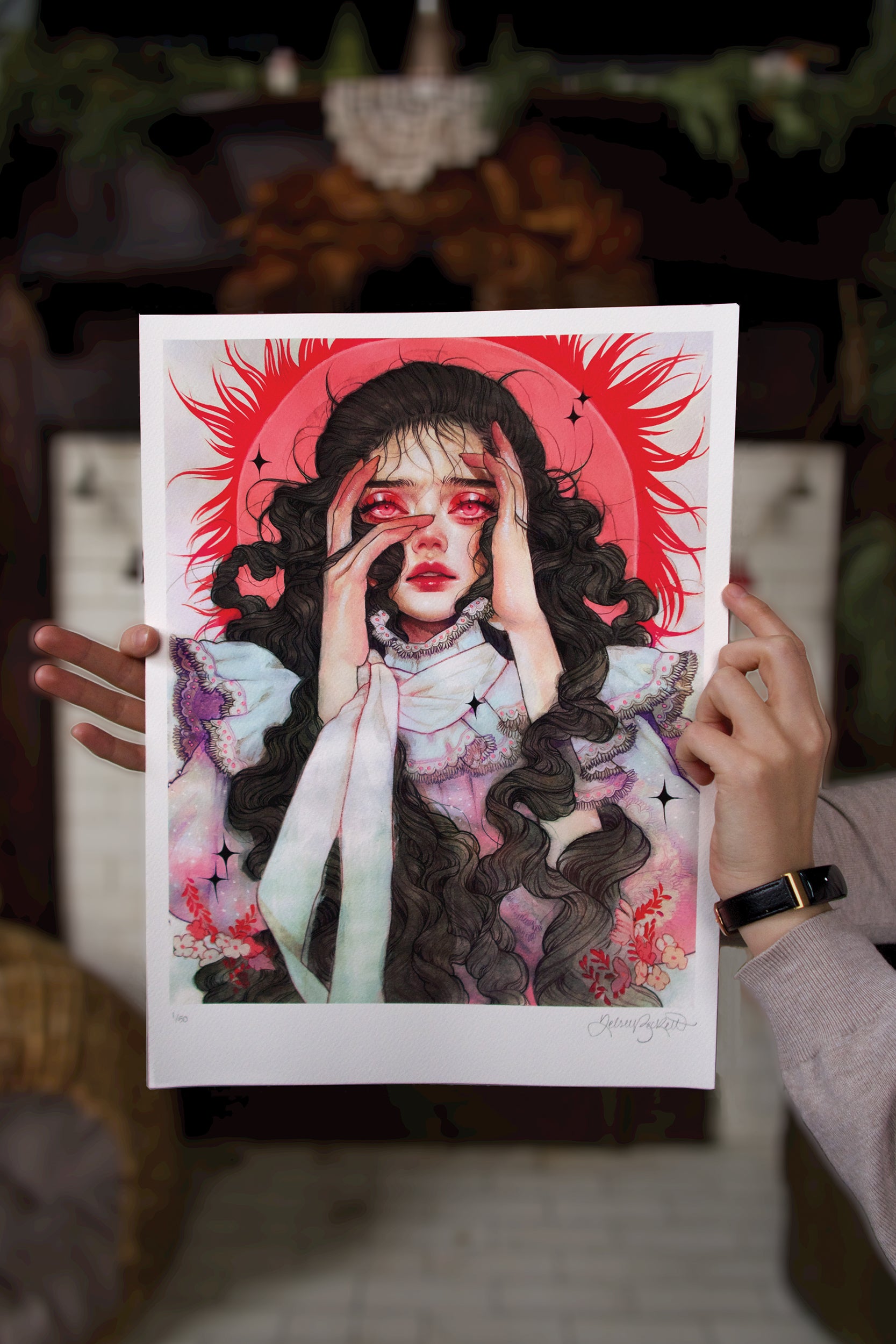 A person holds Dread, a vivid archival pigment print by Kelsey Beckett, showing a woman with long wavy hair against a sun-like red background. The limited edition piece highlights her hands near her face, red accents around her eyes, and is surrounded by floral and abstract elements.