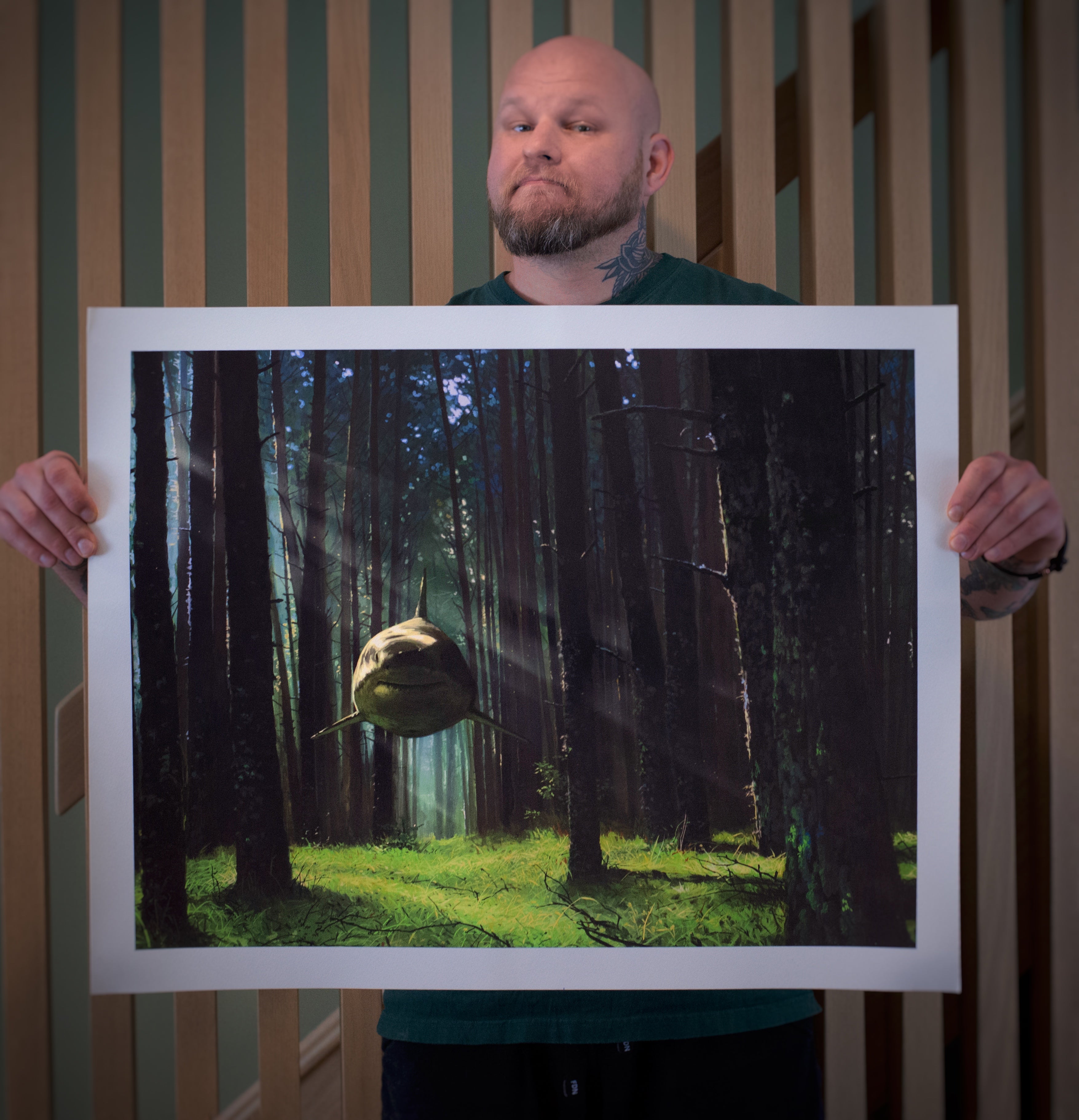 A tattooed individual holds a signed and numbered Ill Hear You Calling Me poster by Chris Austin, showcasing an archival pigment print of a shark swimming through a sunlit forest, beautifully contrasting the ocean creature with the wooded background.