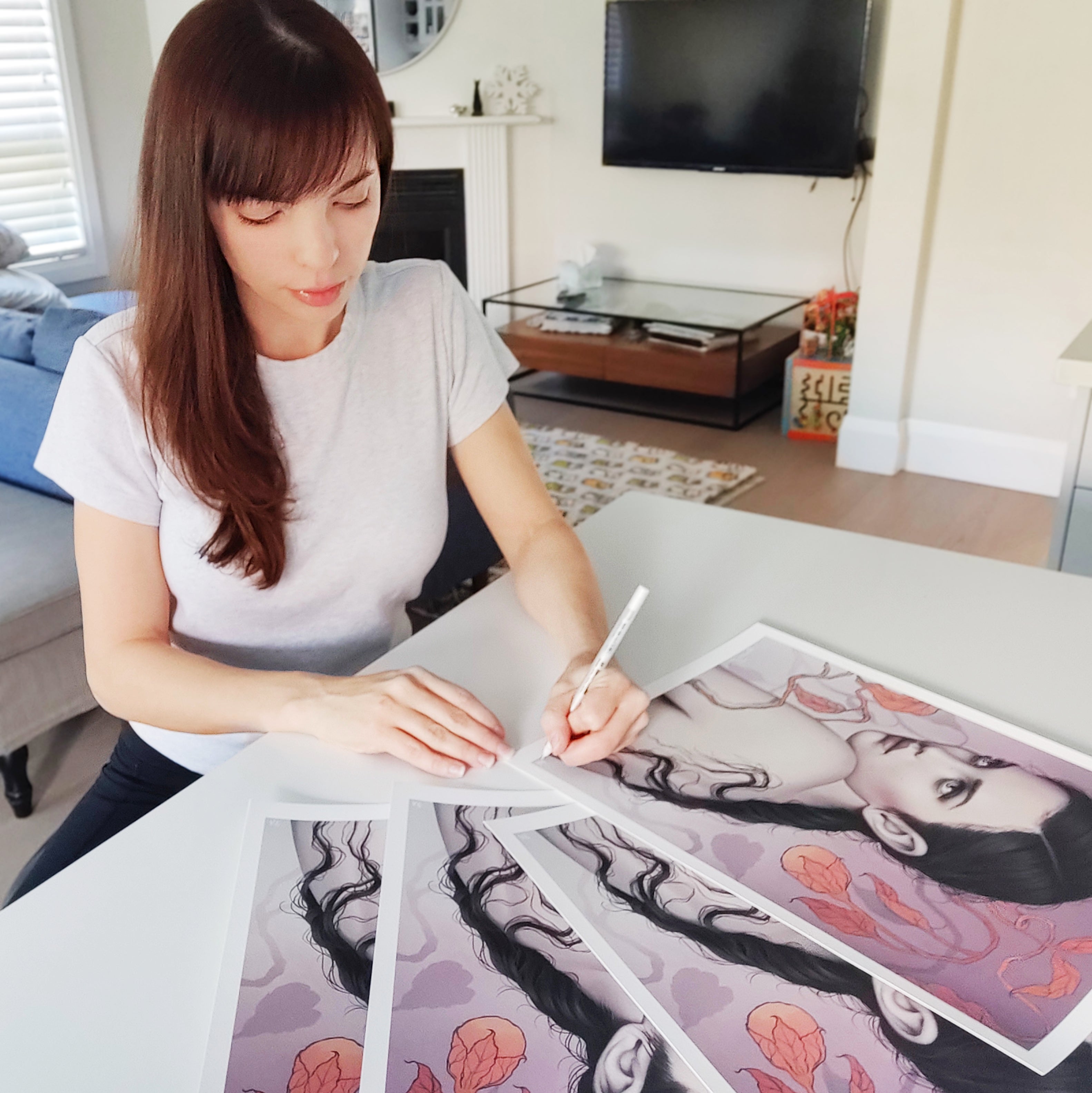 A person with long brown hair, wearing a white shirt, sits in a modern living room signing Sarah Joncas Mind Mischief art prints, which feature a womans portrait with floral elements.
