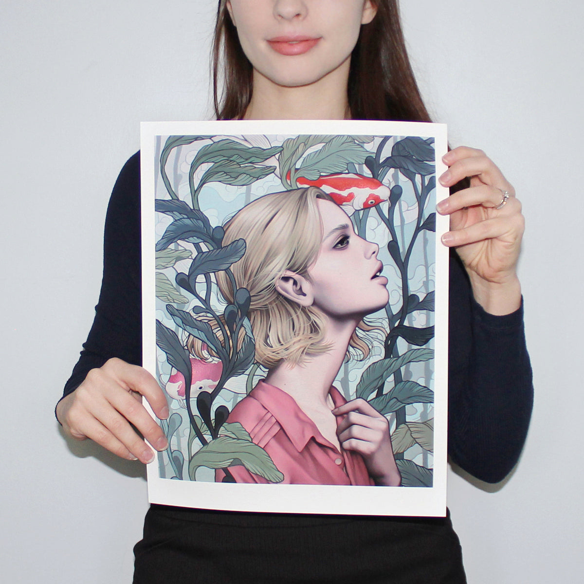 A person holds Sarah Joncas Pond Dreams, an archival pigment print showing a profile of an individual with a blonde bob against a light blue background with circular patterns, surrounded by stylized green leaves and red-orange fish.