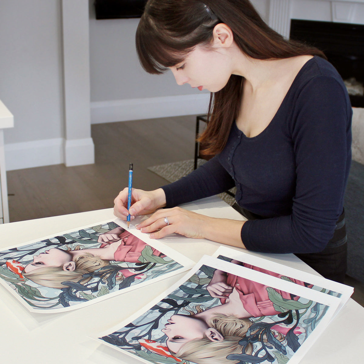 A woman with long brown hair attentively signs Sarah Joncas Archival Pigment Prints, Pond Dreams, at a white table, wearing a dark long-sleeve top. The prints capture the enchanting essence of a girl surrounded by foliage.