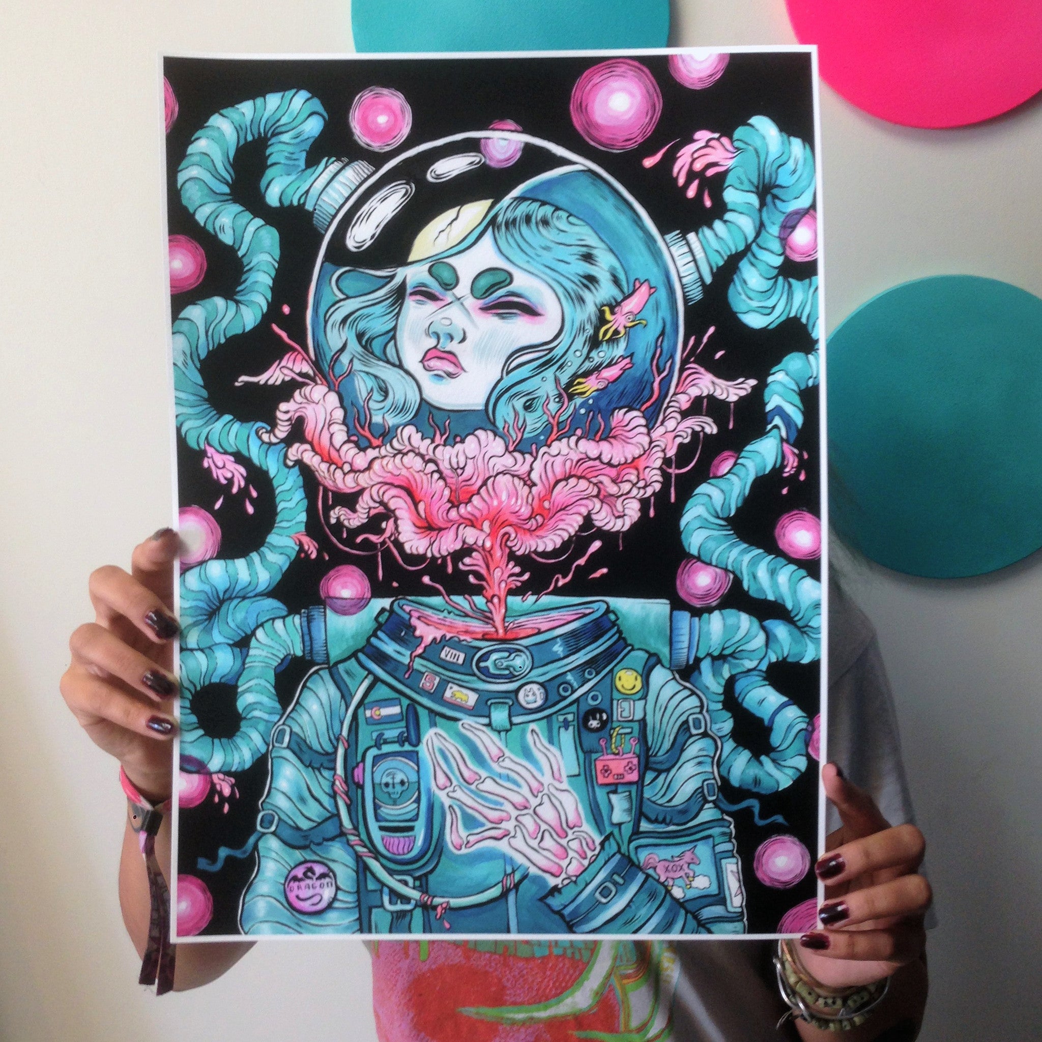 A person holds Growing Up, a vibrant illustration by Lauren YS. This limited edition fine art giclee print depicts an astronaut with nebula-like designs, bright pink and teal circles, and a suit adorned with patches.