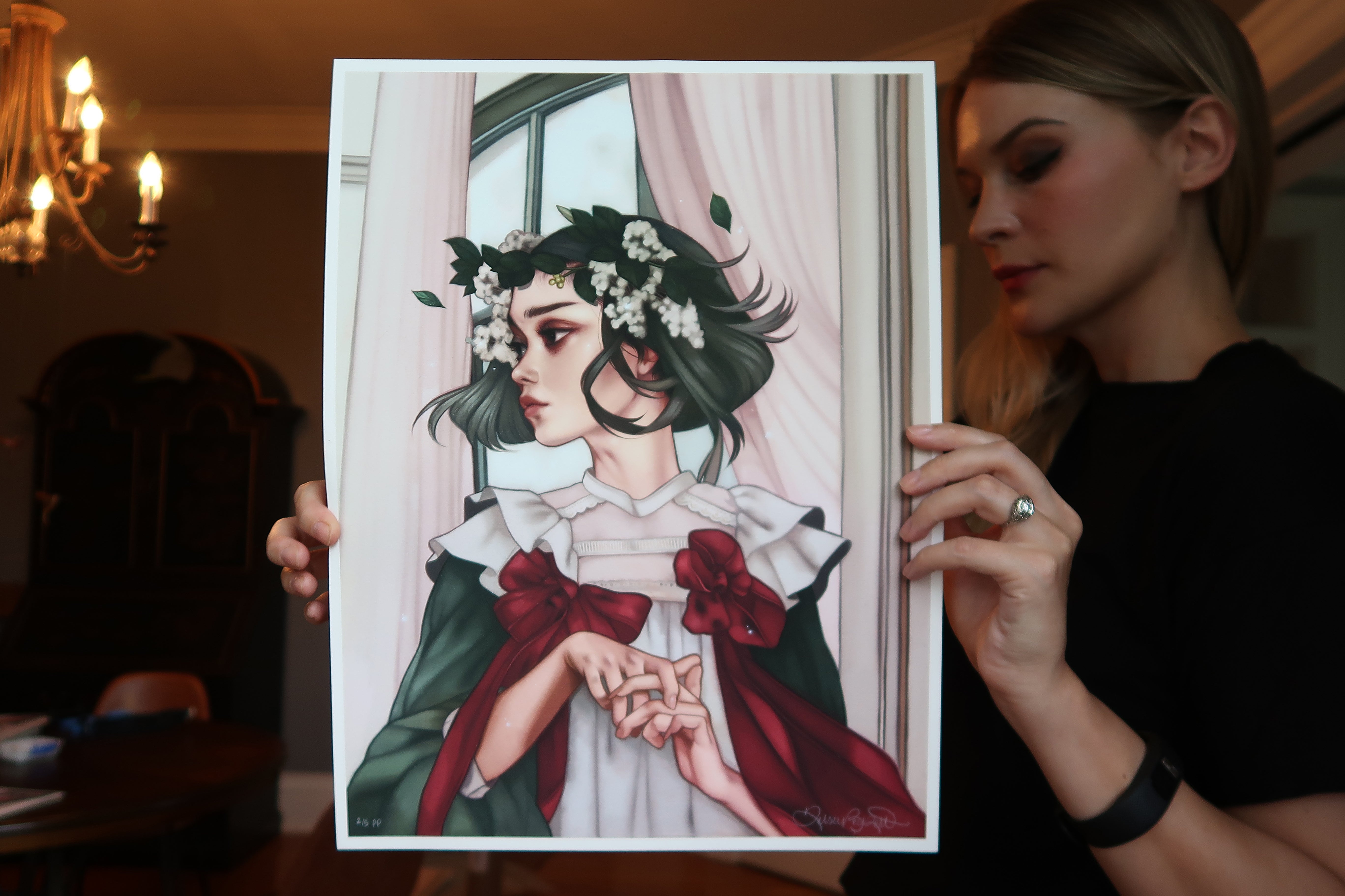 Someone holds Another Last Time, a limited edition print by Kelsey Beckett. It depicts a woman with green hair and white flowers, wearing a red and green dress with a large bow, standing thoughtfully by a window beneath an elegant chandelier.
