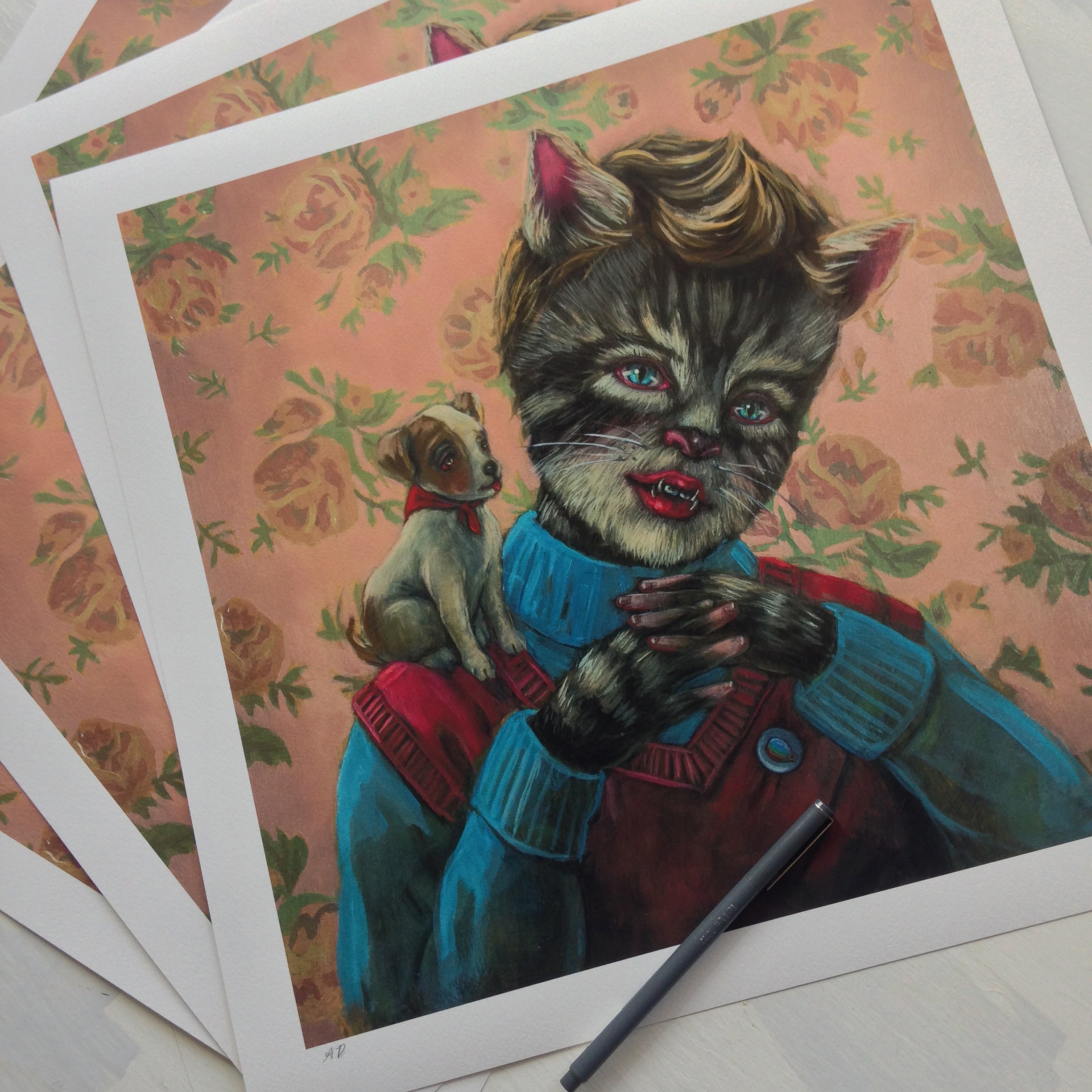 Pussboy" by Lori Nelson depicts a humanoid cat in a red sweater holding a dog on its shoulder, set against florals. Multiple copies of this Archival Pigment Print are stacked beneath, with a pen resting on the white border.