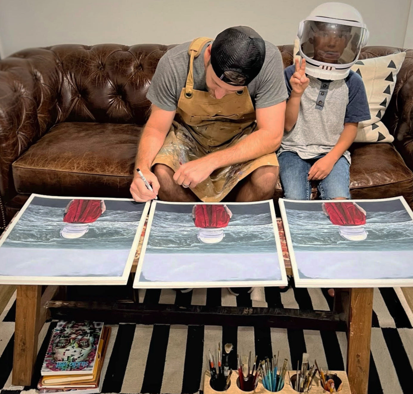 An individual in a cap and apron sits on a couch, signing and numbering archival pigment prints of the Adam Hall artwork A Presence of Wonder. Beside them, a child in a space helmet flashes a peace sign amidst scattered art supplies on the carpeted floor.