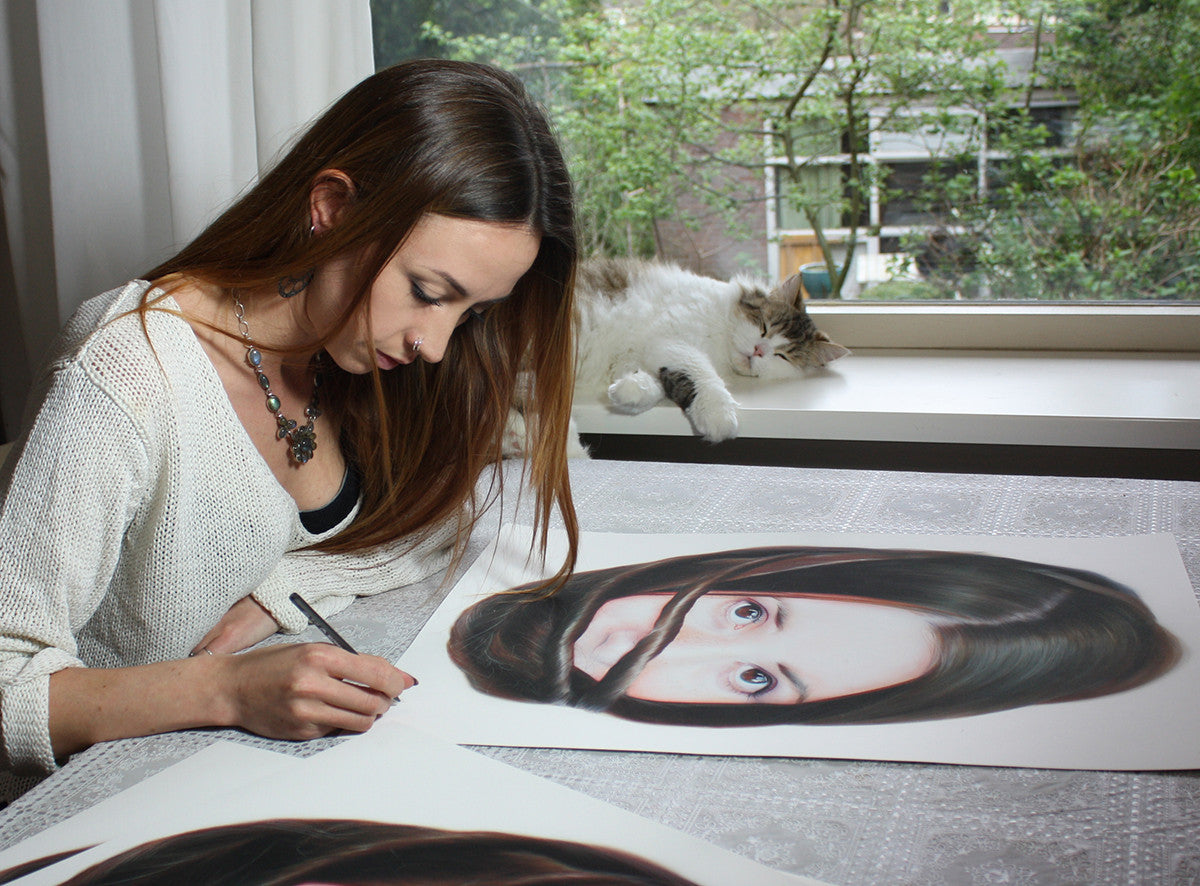 A woman with long brown hair sketches a portrait for Roos van der Vliets Storytellers XV series at a table. A fluffy cat lounges by the window, which overlooks lush green trees, making the scene resemble a vivid archival pigment print.