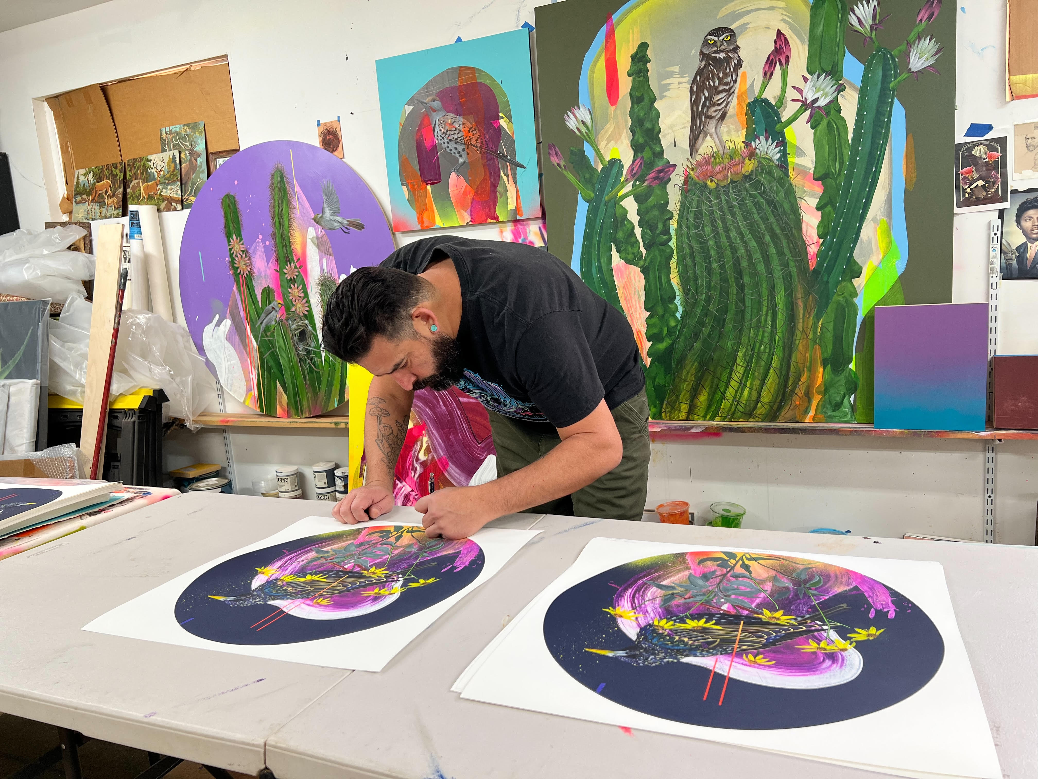 Artist Frank Gonzales is creating a colorful artwork titled Shakespeare Burden at a studio table, surrounded by his vibrant paintings of cacti and animals. The lively scene includes numerous signed and numbered archival pigment prints.