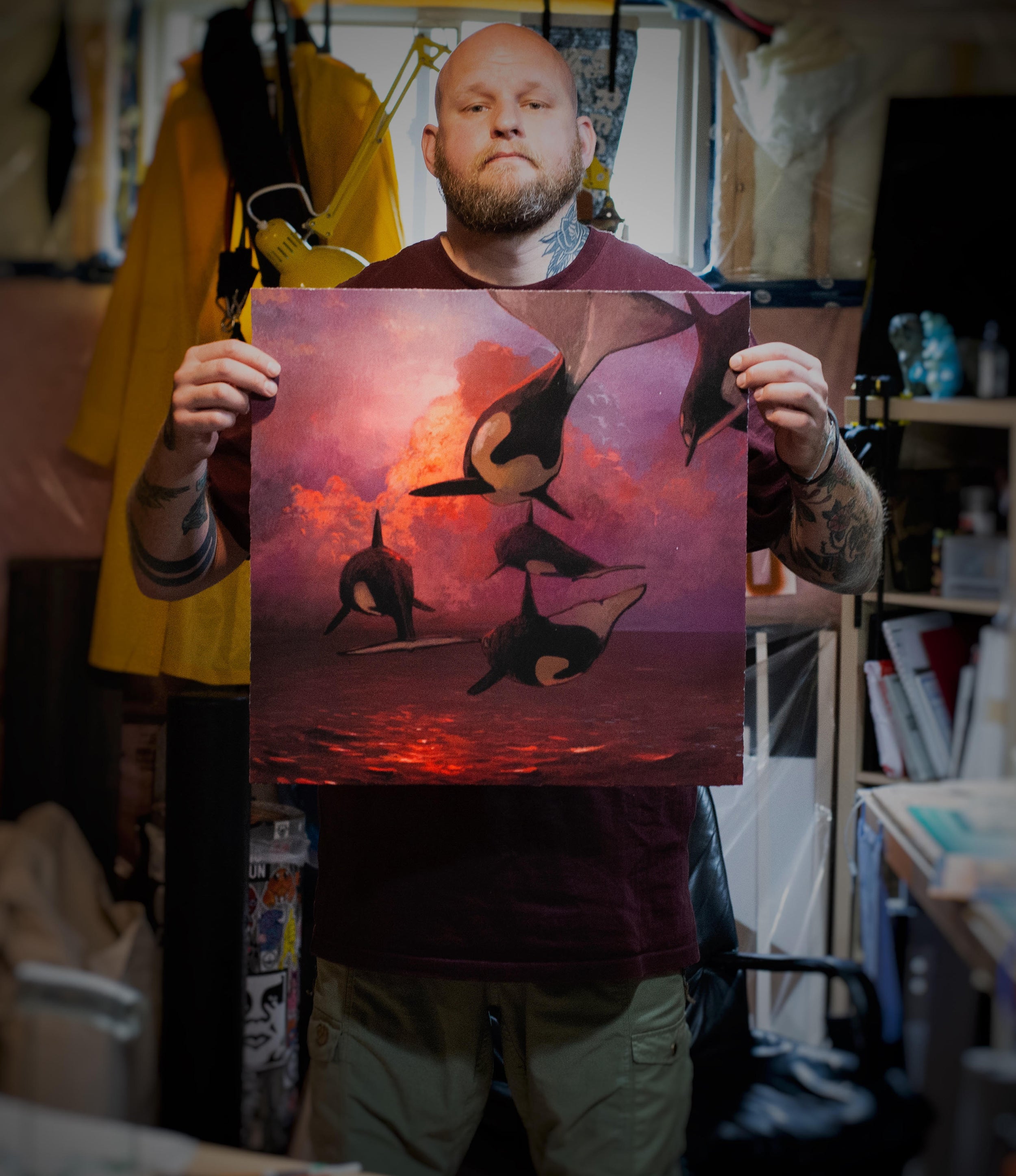 Indoors, a bearded person holds Where the Sky Touches the Sea, a vivid archival pigment print by Chris Austin. The artwork features orcas leaping through a reddish-purple ocean against a dramatic sky. Part of a hand-embellished edition, it beautifully showcases multiple colors and marine themes.