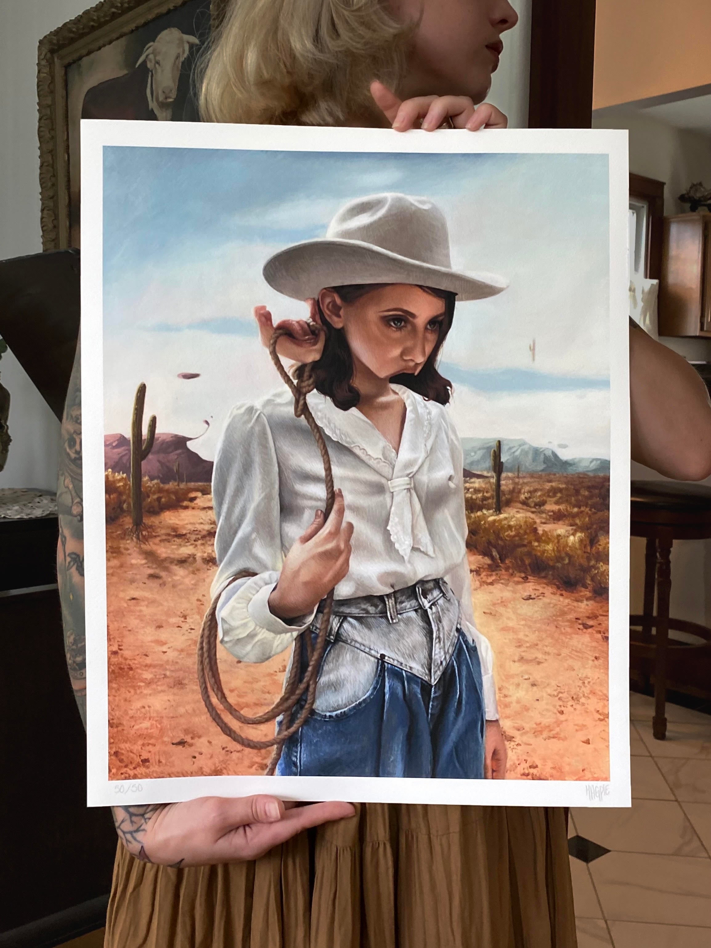 A person holds the Meagan Magpie Rodgers archival pigment print Alone With My Thoughts, depicting a woman in a cowboy hat and white shirt in a desert landscape with cacti, with furniture and wall decor indoors.