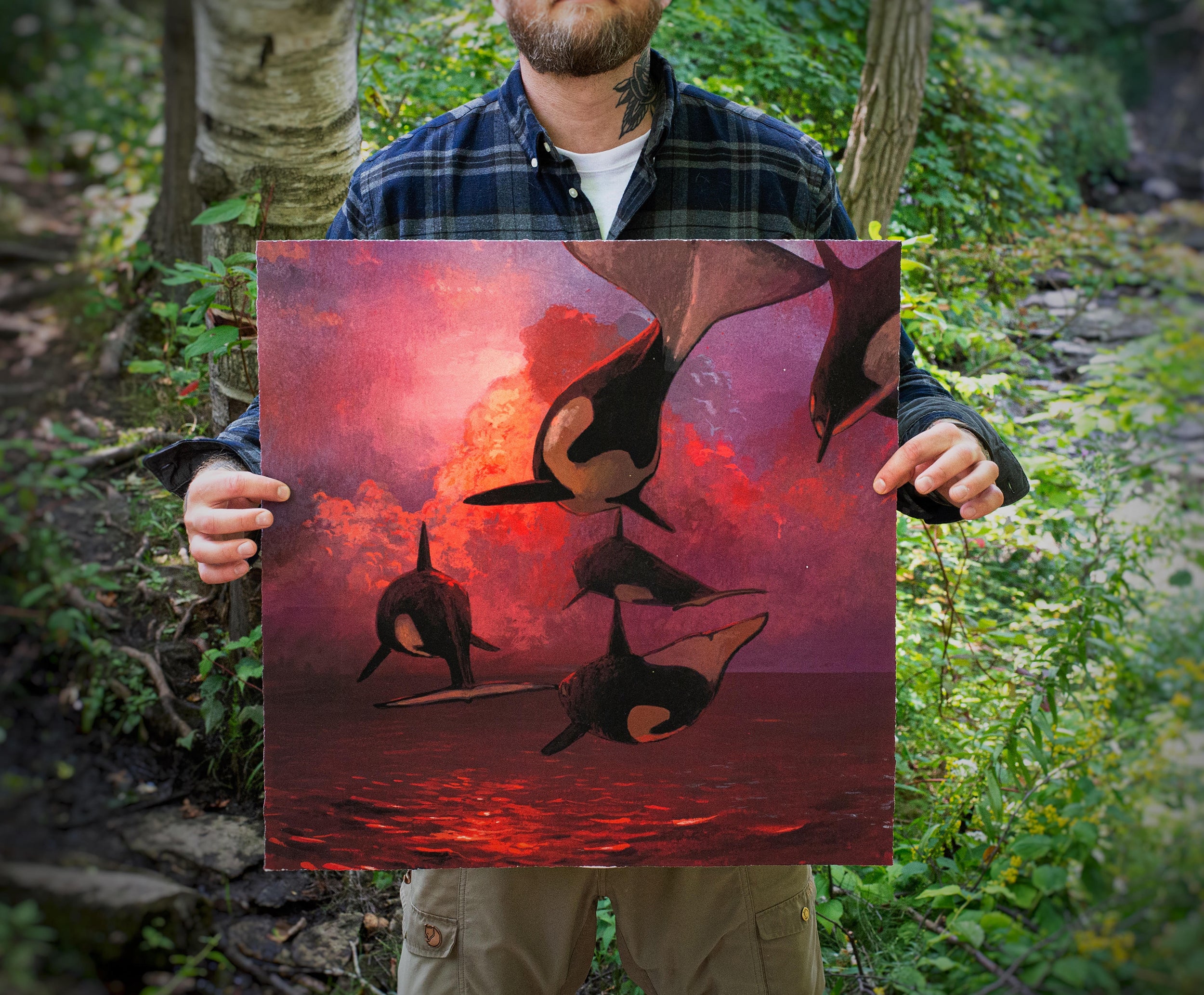 A person holds Chris Austins Where the Sky Touches the Sea, a striking archival pigment print of orcas against a vivid sunset. Set in a lush forest, the individual wears a plaid shirt and sports a neck tattoo, mirroring nature’s raw beauty.