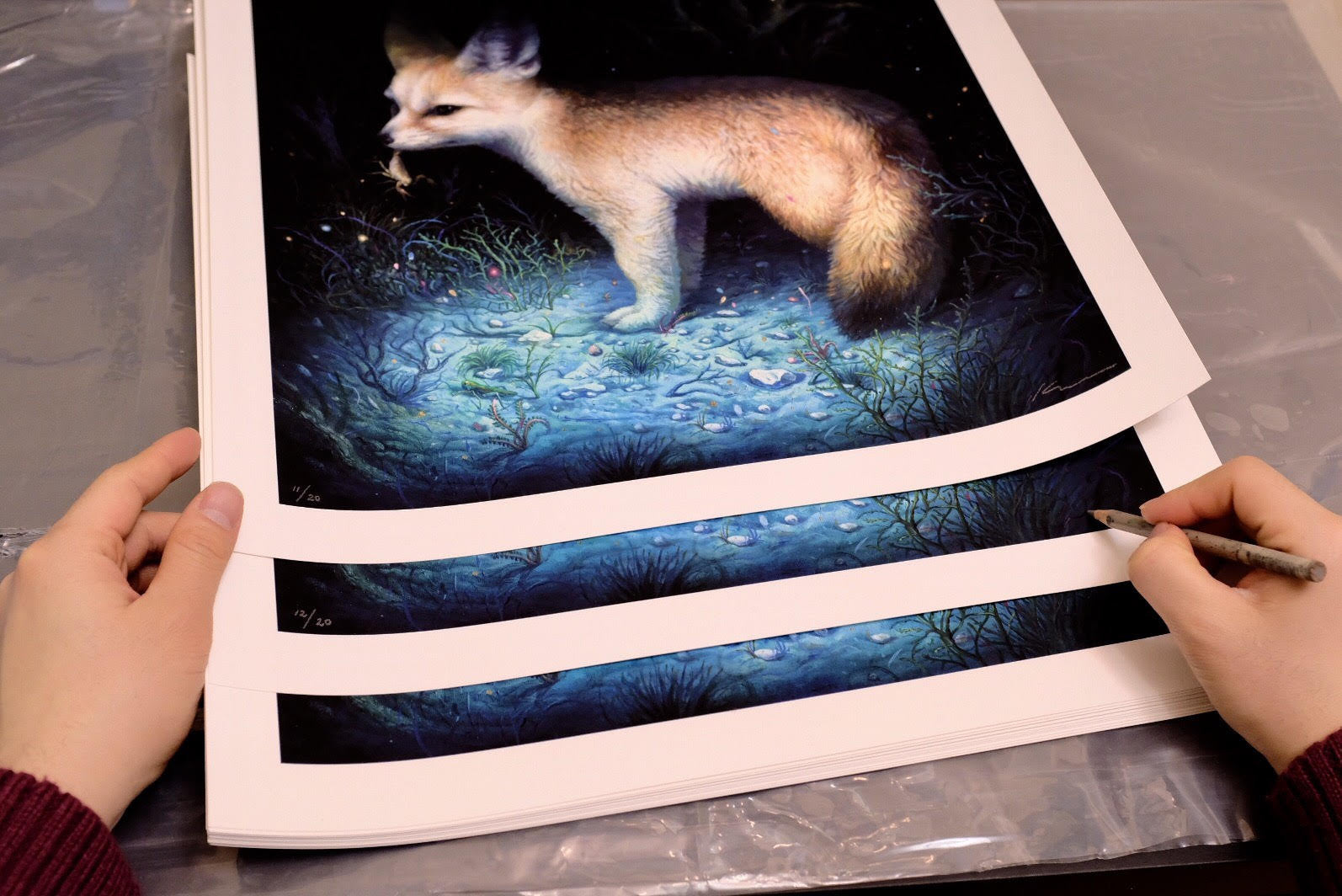 A person holds KiSung Koh's "Neith" art prints featuring a fox on a glowing blue forest floor. The signed, numbered edition is autographed by the artist in pencil on the bottom corner as the prints rest on plastic.