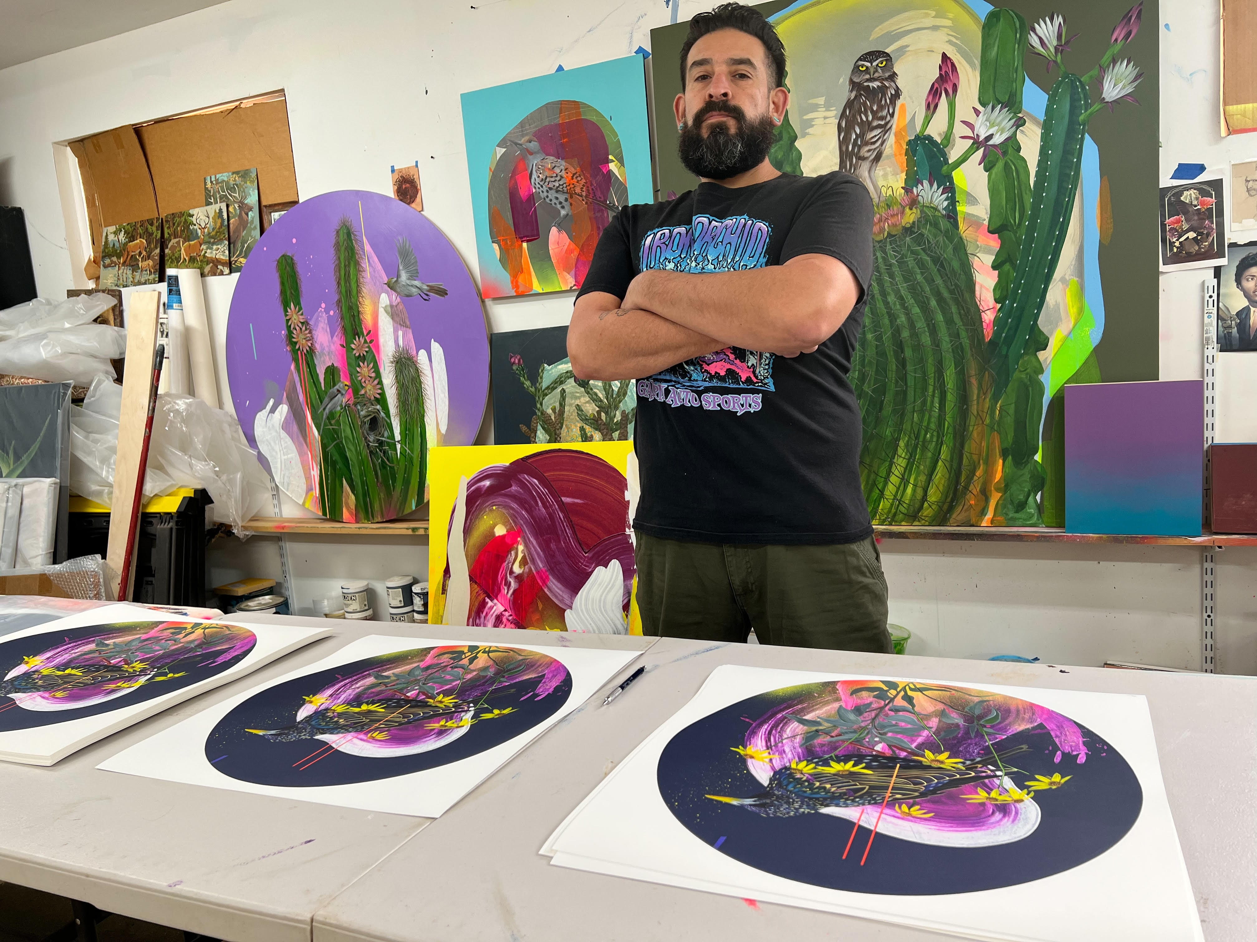 In Frank Gonzaless studio, an artist with a beard and crossed arms is surrounded by colorful artwork titled Shakespeare Burden. These vibrant archival pigment prints feature signed and numbered nature themes with plants, birds, and abstract designs, displayed on tables and walls.