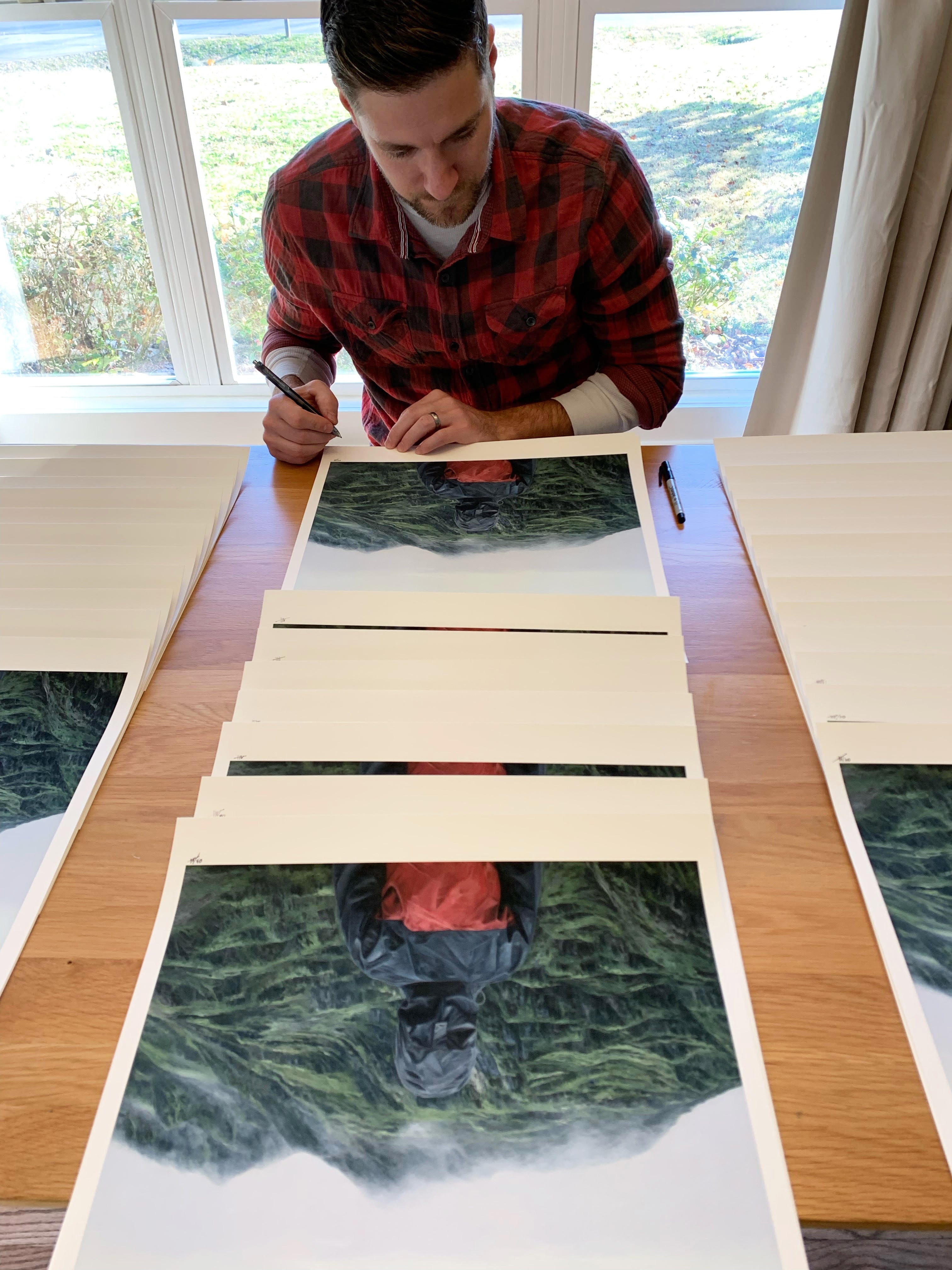 Dressed in a red plaid shirt, someone signs and numbers each Acclimation archival pigment print from an edition of 50. The Adam Hall art prints, depicting a figure in a raincoat in misty mountains, lie on a wooden table illuminated by sunlight streaming through the window.