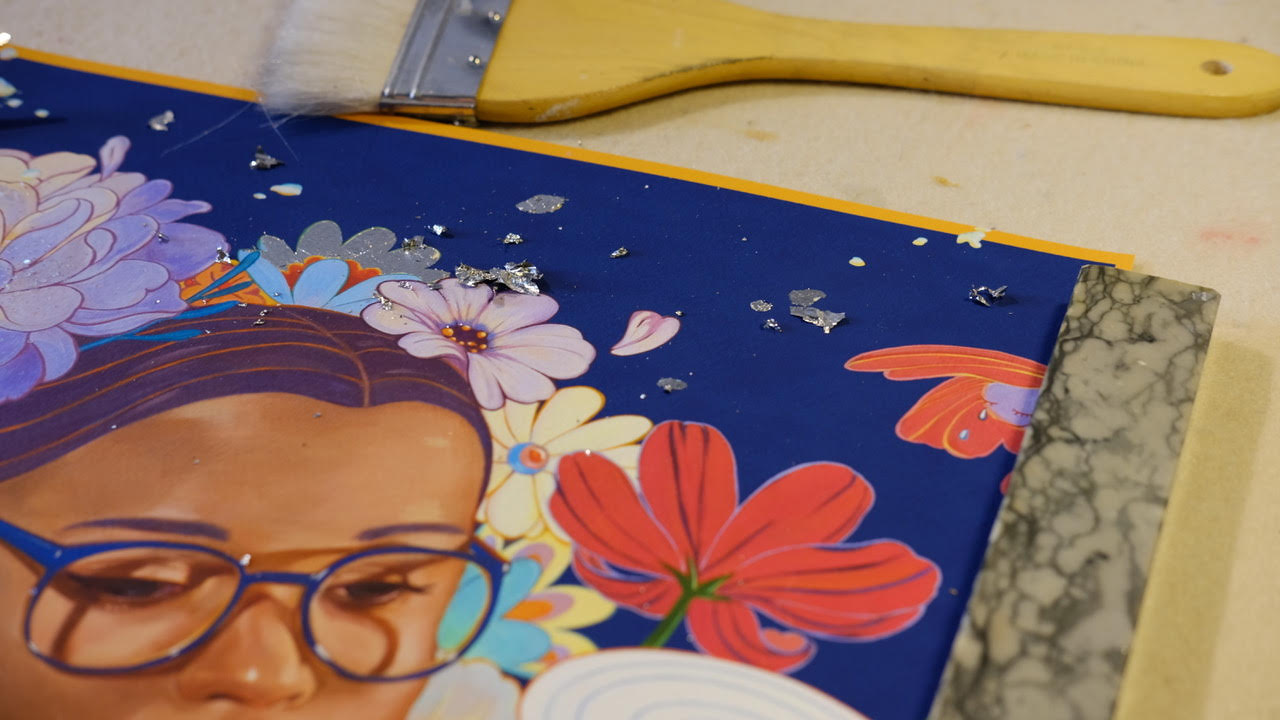Sweetness No. 5 by Helice Wen showcases a close-up artistic piece of a person with glasses and floral motifs on a blue background, adorned with silver leaf. Nearby, an archival pigment print is meticulously hand-embellished next to a paintbrush on the table.
