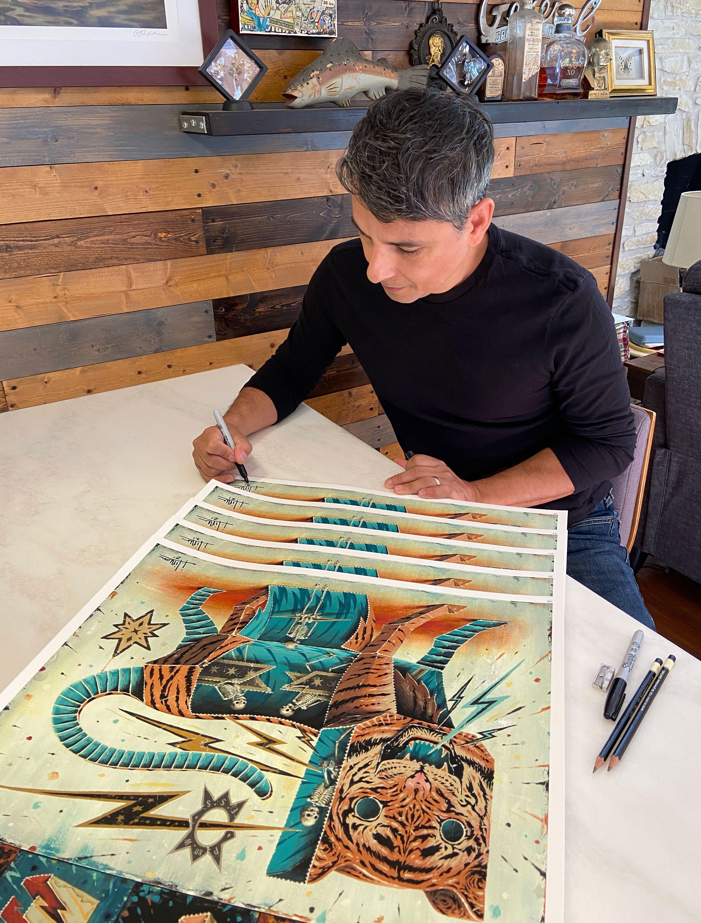 A person with short dark hair sits at a table, signing and numbering Prowess, an archival pigment print by Jason Limon featuring a stylized tiger with vibrant geometric patterns against a wood-paneled wall filled with assorted framed items.
