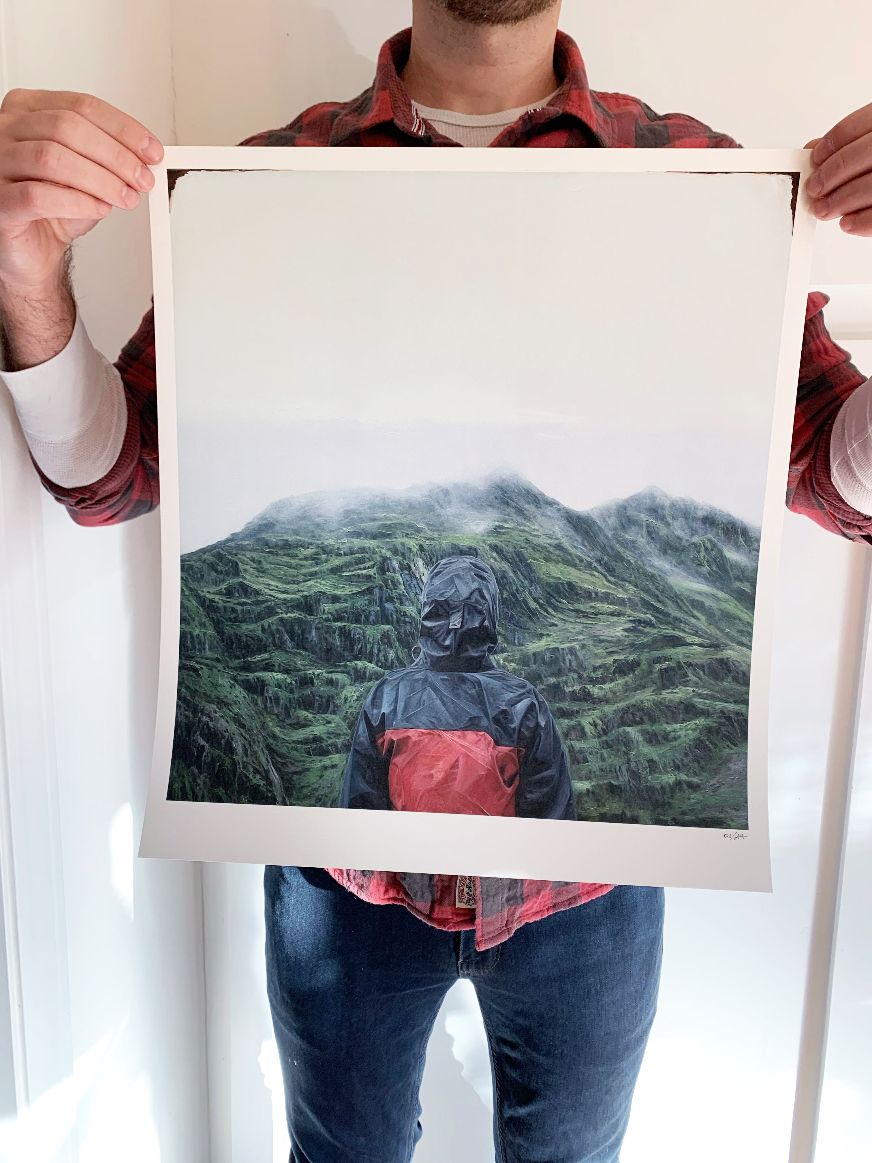A person wearing a red and black jacket holds Acclimation, a large signed and numbered photo print by Adam Hall, featuring a figure in a similar jacket overlooking a foggy green mountainous landscape.