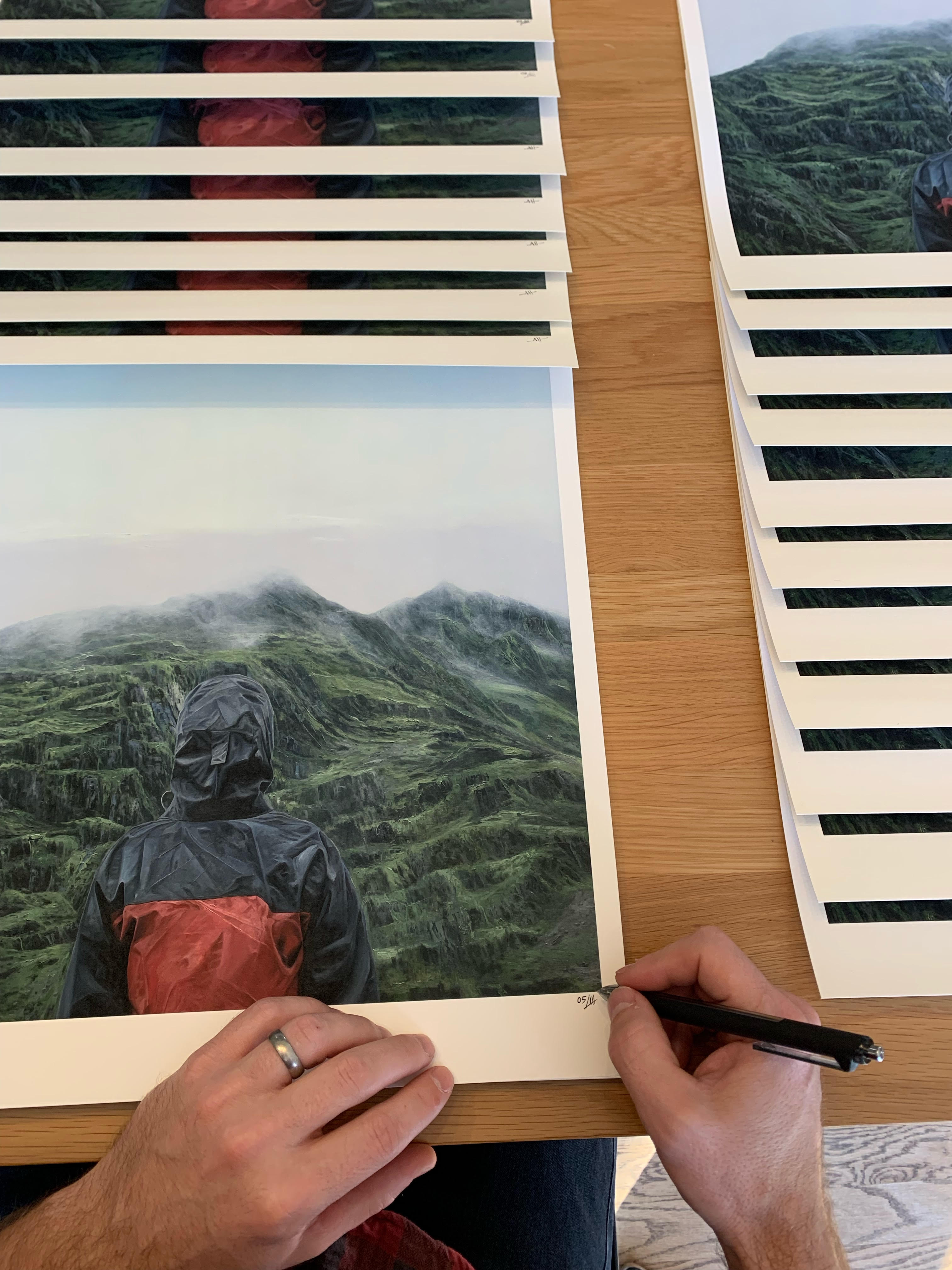 Adam Halls limited edition print Acclimation features 50 archival pigment prints, each showing a figure in a red and black jacket before misty green mountains. These signed and numbered pieces are neatly stacked on a wooden table.