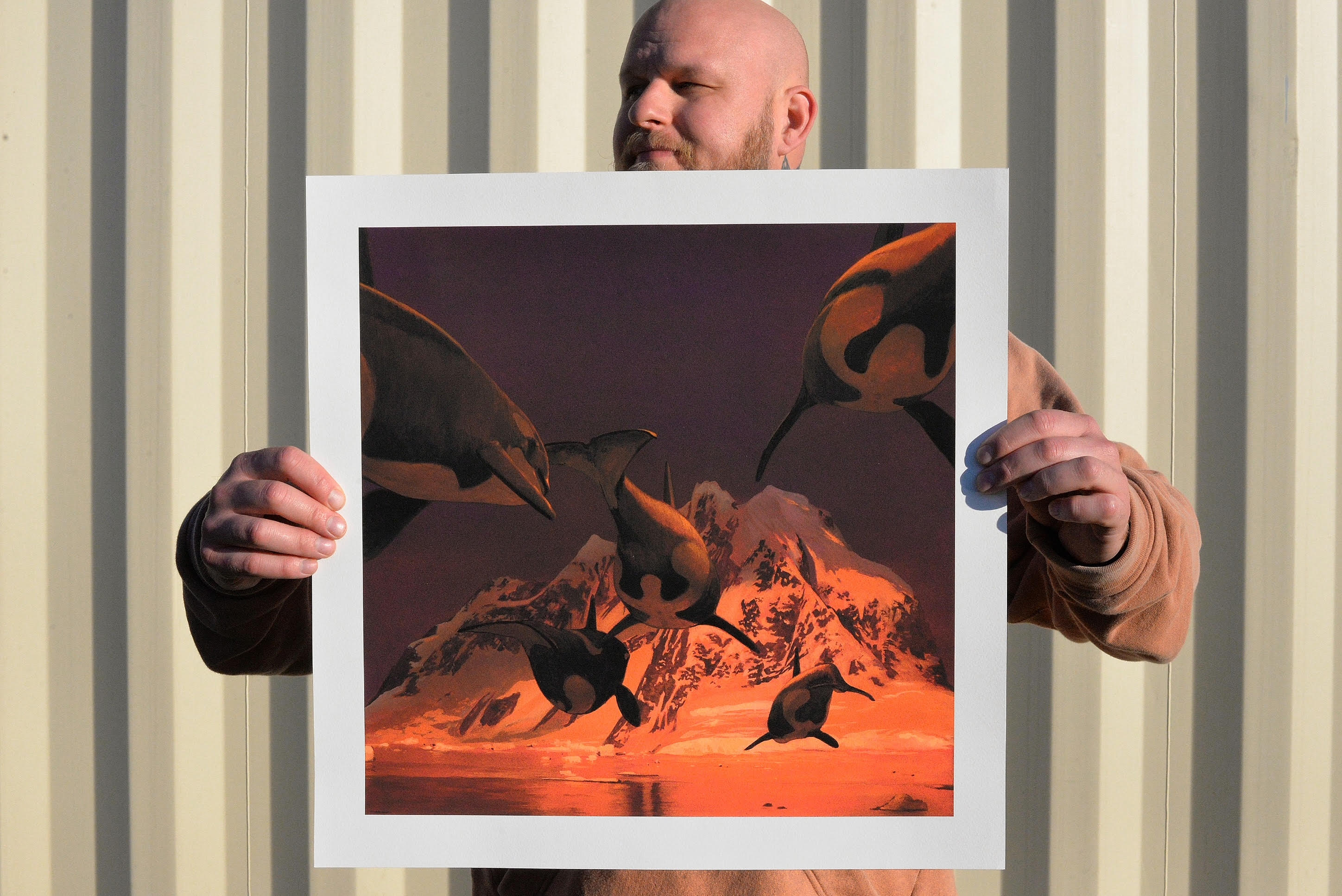 A person holds In A Silent Way, a signed and numbered art print by Chris Austin, showcasing orcas swimming against mountain backdrops with reddish tones and vivid contrasts, set against a corrugated metal wall.