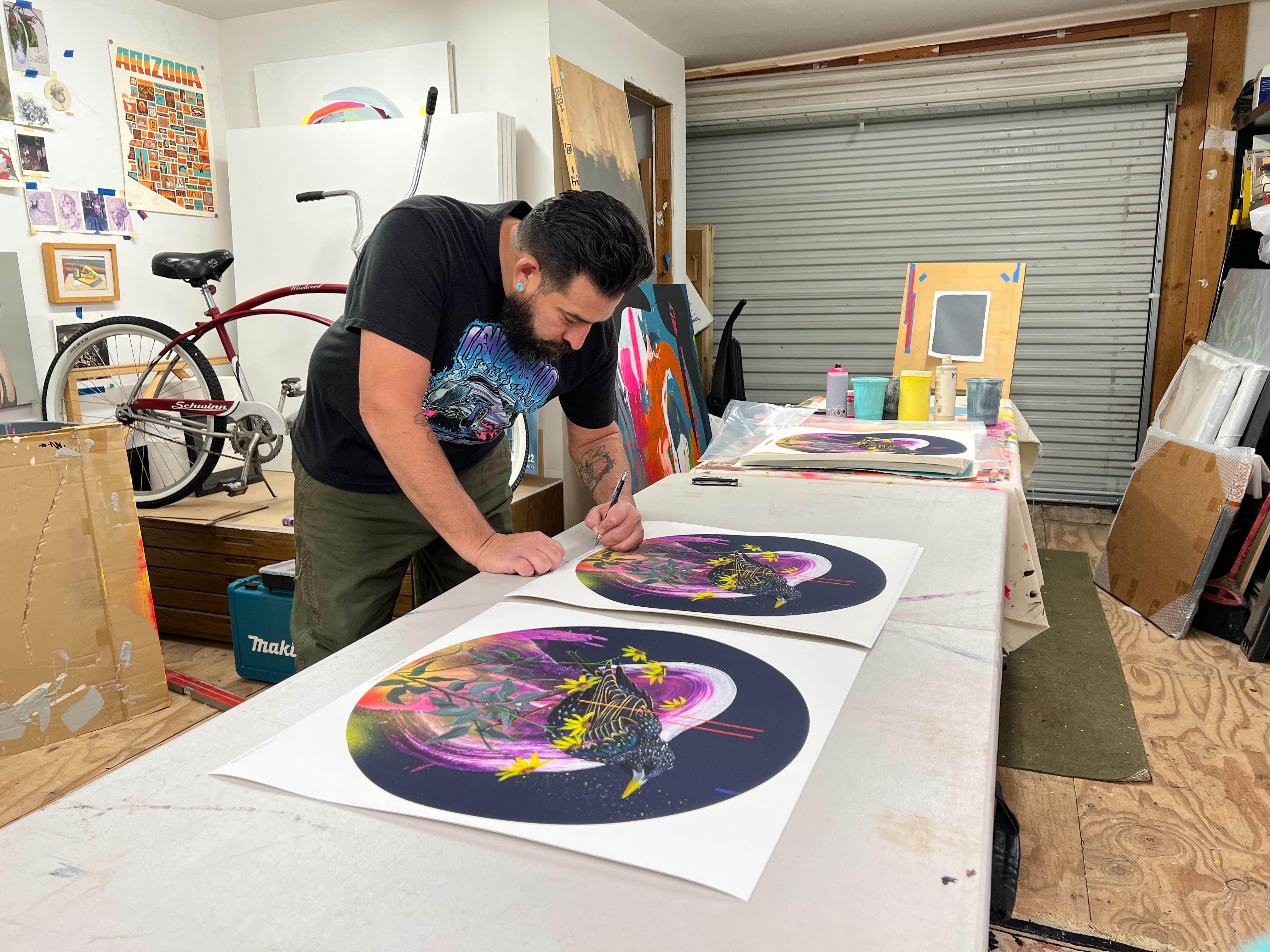 In an art studio filled with supplies and finished artworks, a bearded person paints colorful circular pieces on a table. A red bicycle sits in the background, alongside a signed and numbered Shakespeare Burden by Frank Gonzales.